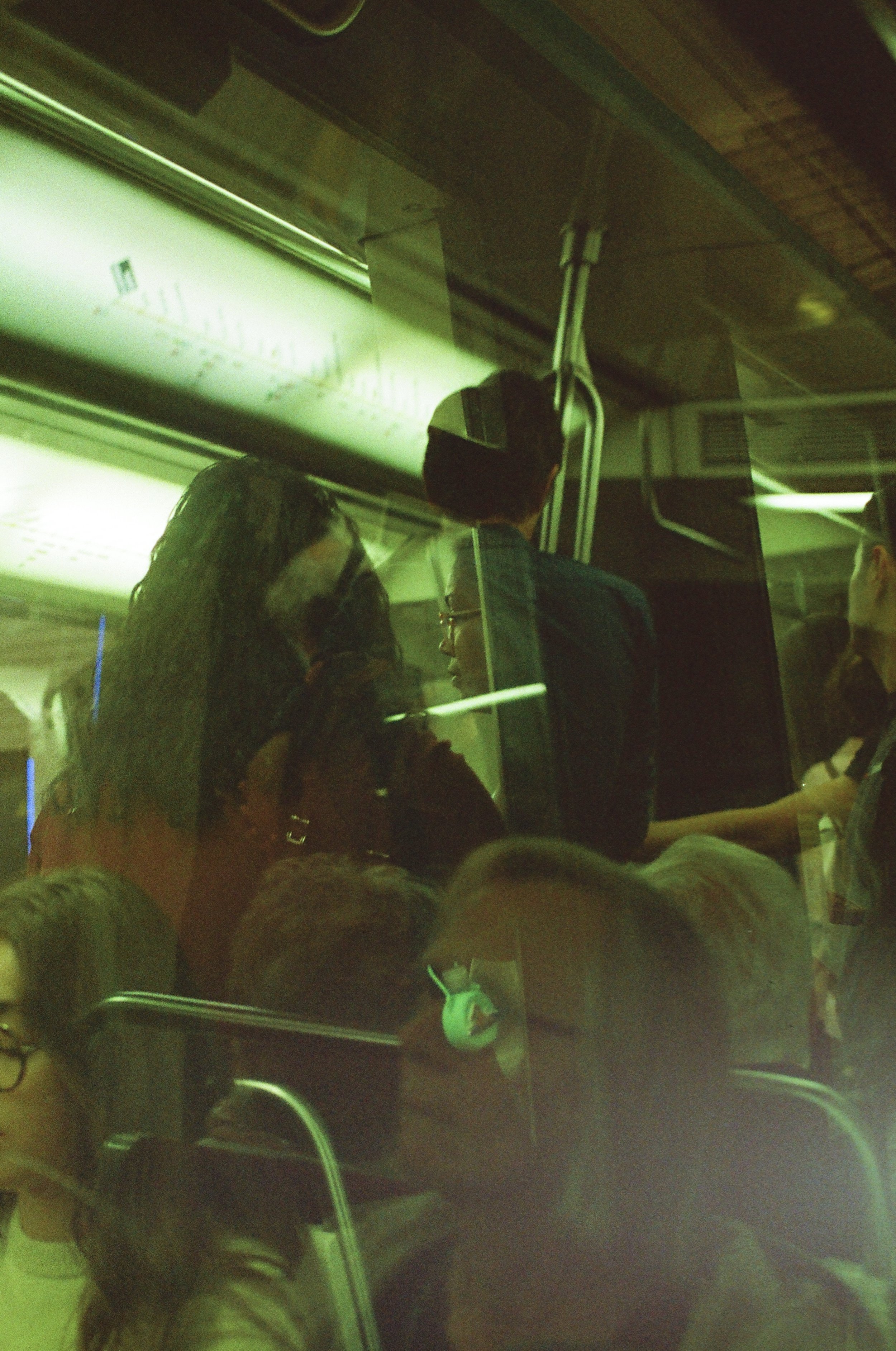 Passengers seated inside a green-lit subway train, with some standing and engaged in conversation, seen through the train window.