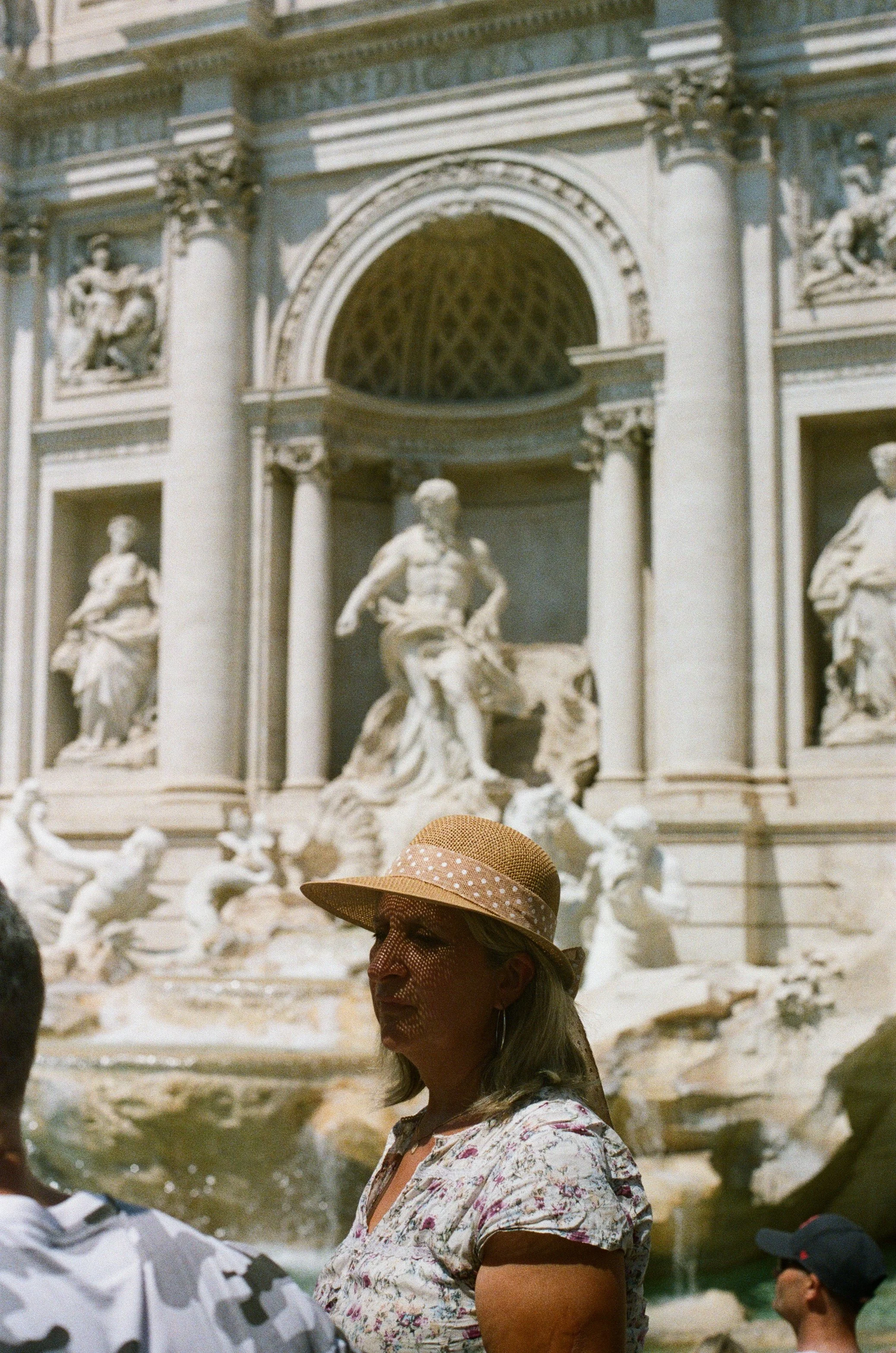 A woman wearing a floral top and a wide-brimmed straw hat with a polka dot ribbon stands in front of the Trevi Fountain in Rome, Italy, with other tourists nearby.