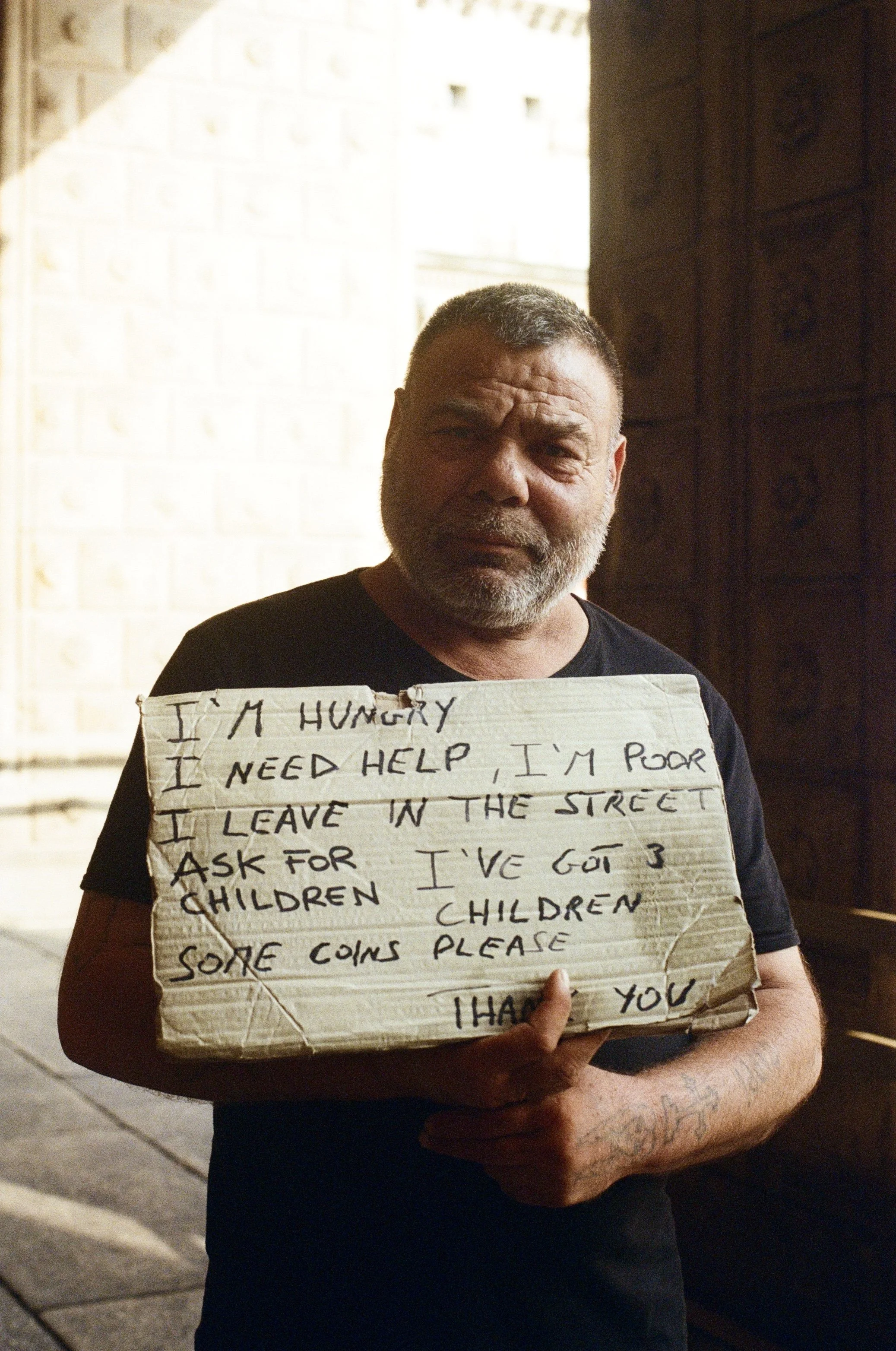 A man with a beard holding a cardboard sign that reads, "I'M HUNGRY, I NEED HELP, I LEAVE IN THE STREET, ASK FOR CHILDREN, SOME COINS PLEASE, THANK YOU."