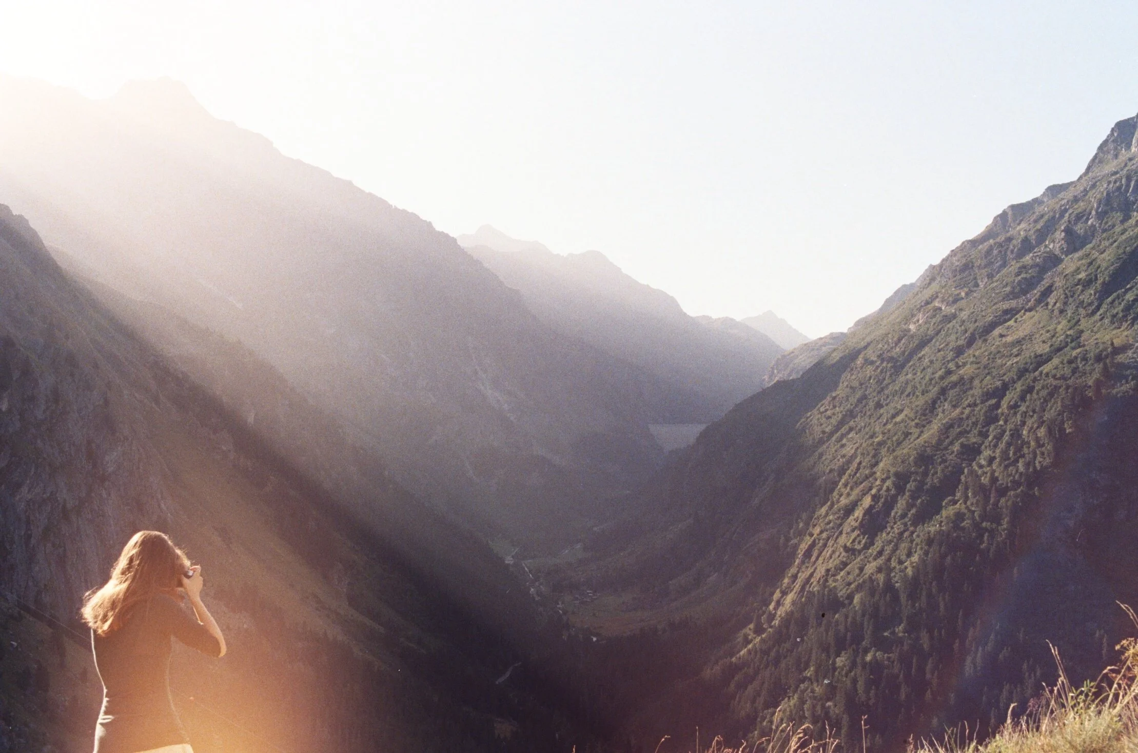 A woman with long hair taking a photo of a scenic mountain valley with sunlight streaming over the peaks.