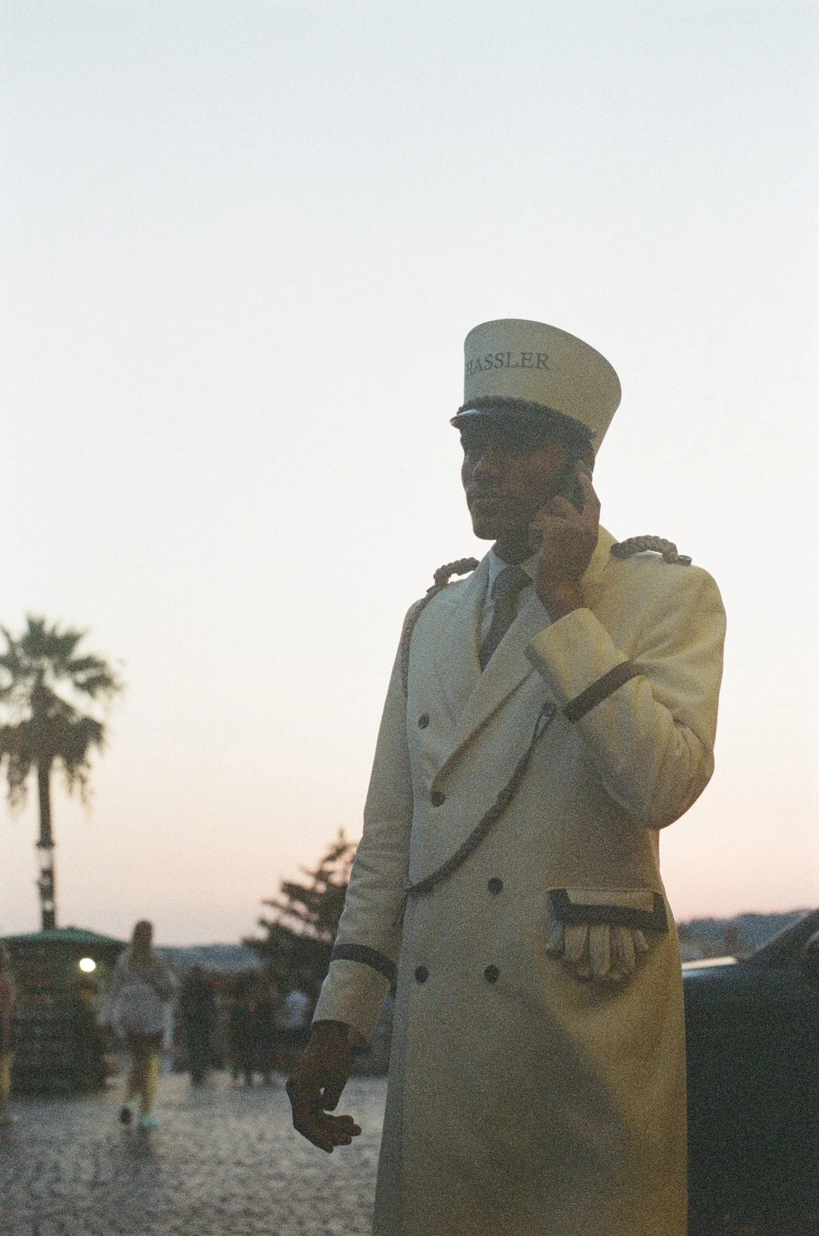 A person wearing a white conductor's outfit and hat with the word 'BASSLER' on it, talking on a cellphone outdoors during sunset, with palm trees and people walking in the background.