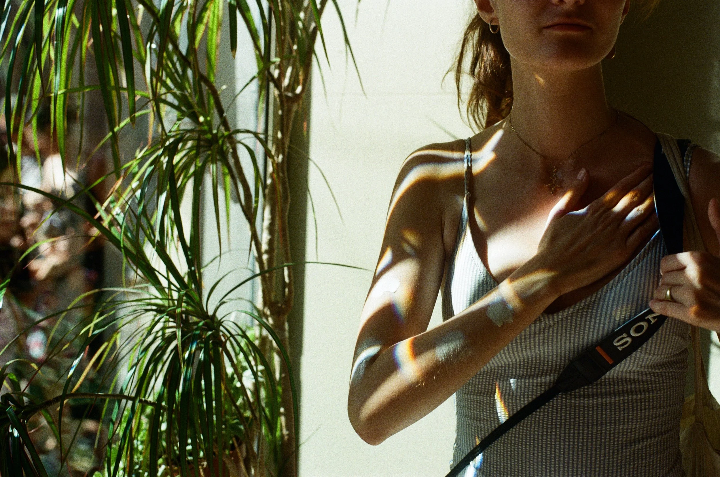 A woman standing near a plant with sunlight creating rainbow-like patterns on her skin, wearing a tank top, a necklace, and a camera strap over her shoulder.
