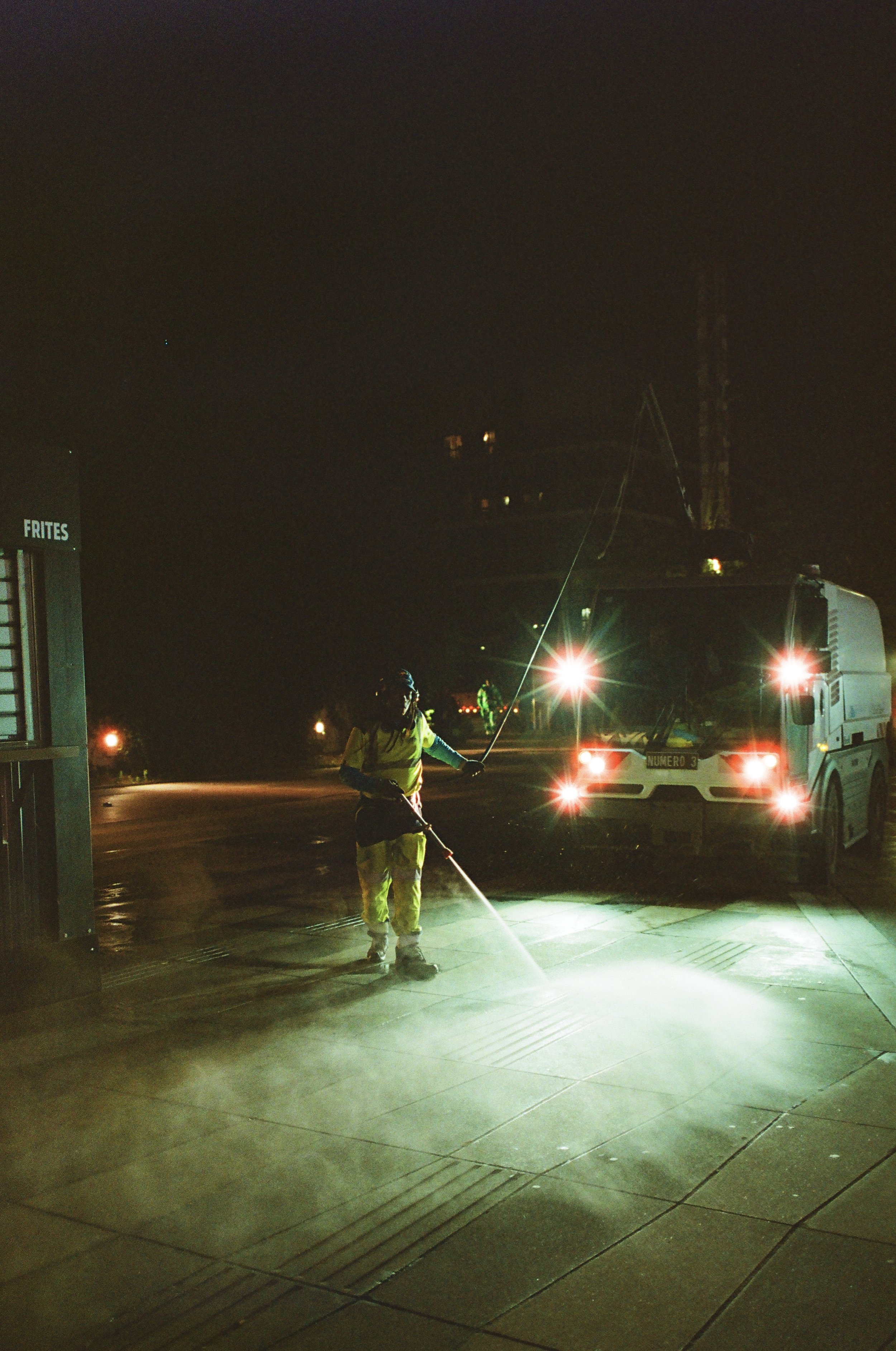 Nighttime scene of a worker cleaning the sidewalk with a high-pressure water hose, illuminated by streetlights and vehicle headlights.