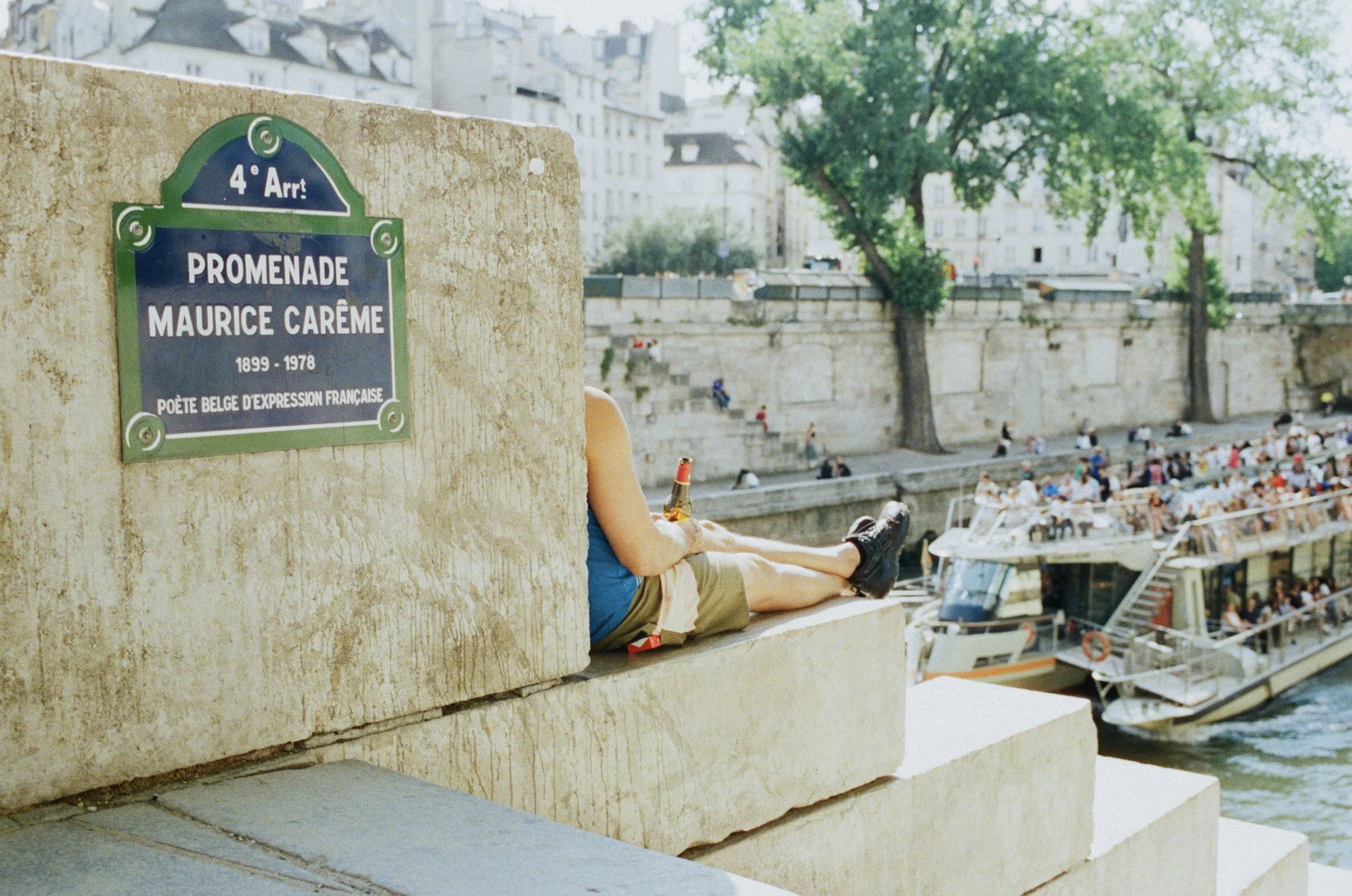 A person sitting on a stone ledge near a river, holding a drink, with a large boat and people walking along the riverbank in the background on a sunny day.