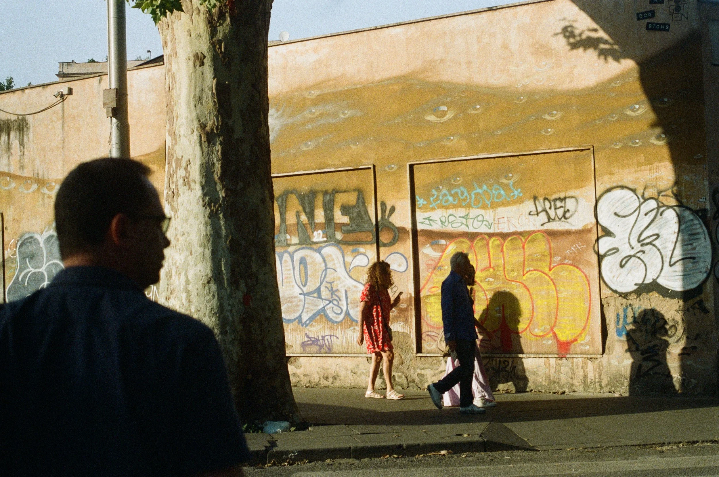 Three people walking past a graffitied wall with a tree in the foreground and shadows cast on the wall.