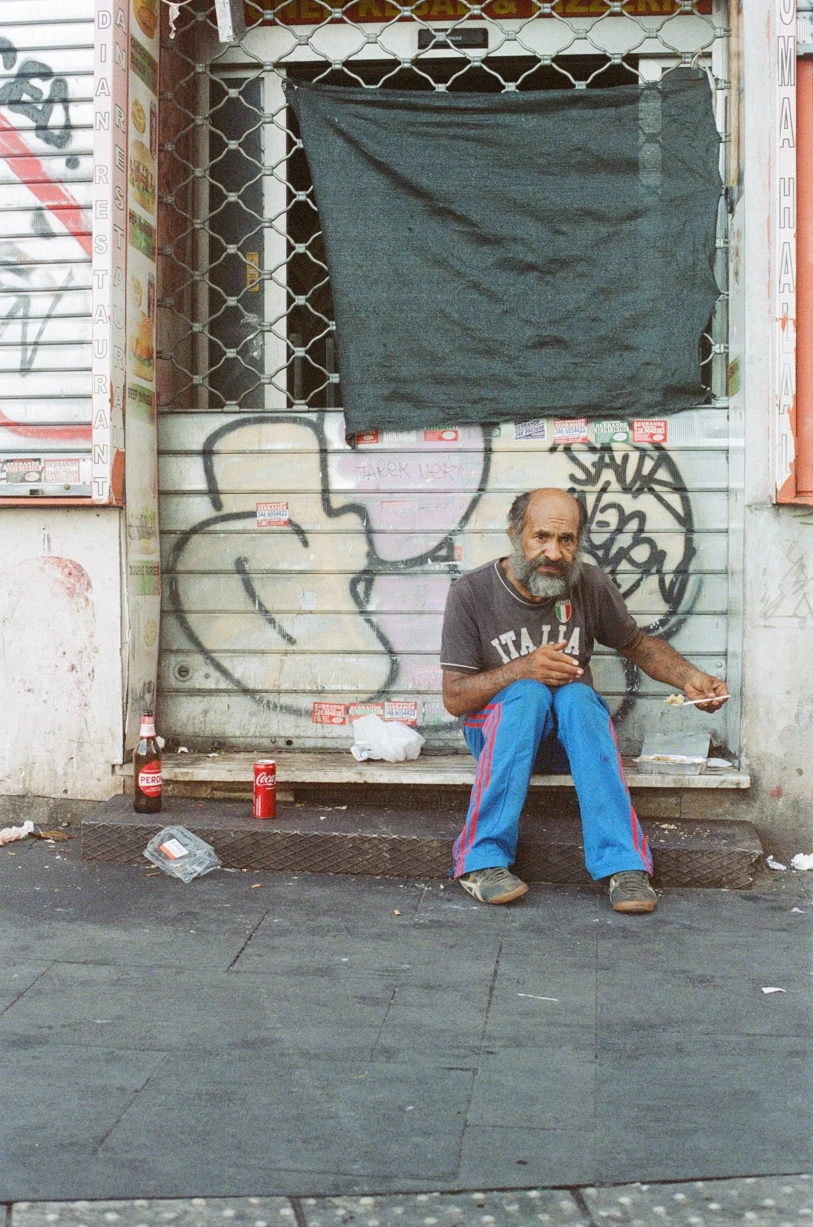 An older man with a beard sitting on a sidewalk in front of a graffitied wall, eating food with a fork, with a bottle of Peroni beer, a soda can, and some trash nearby.