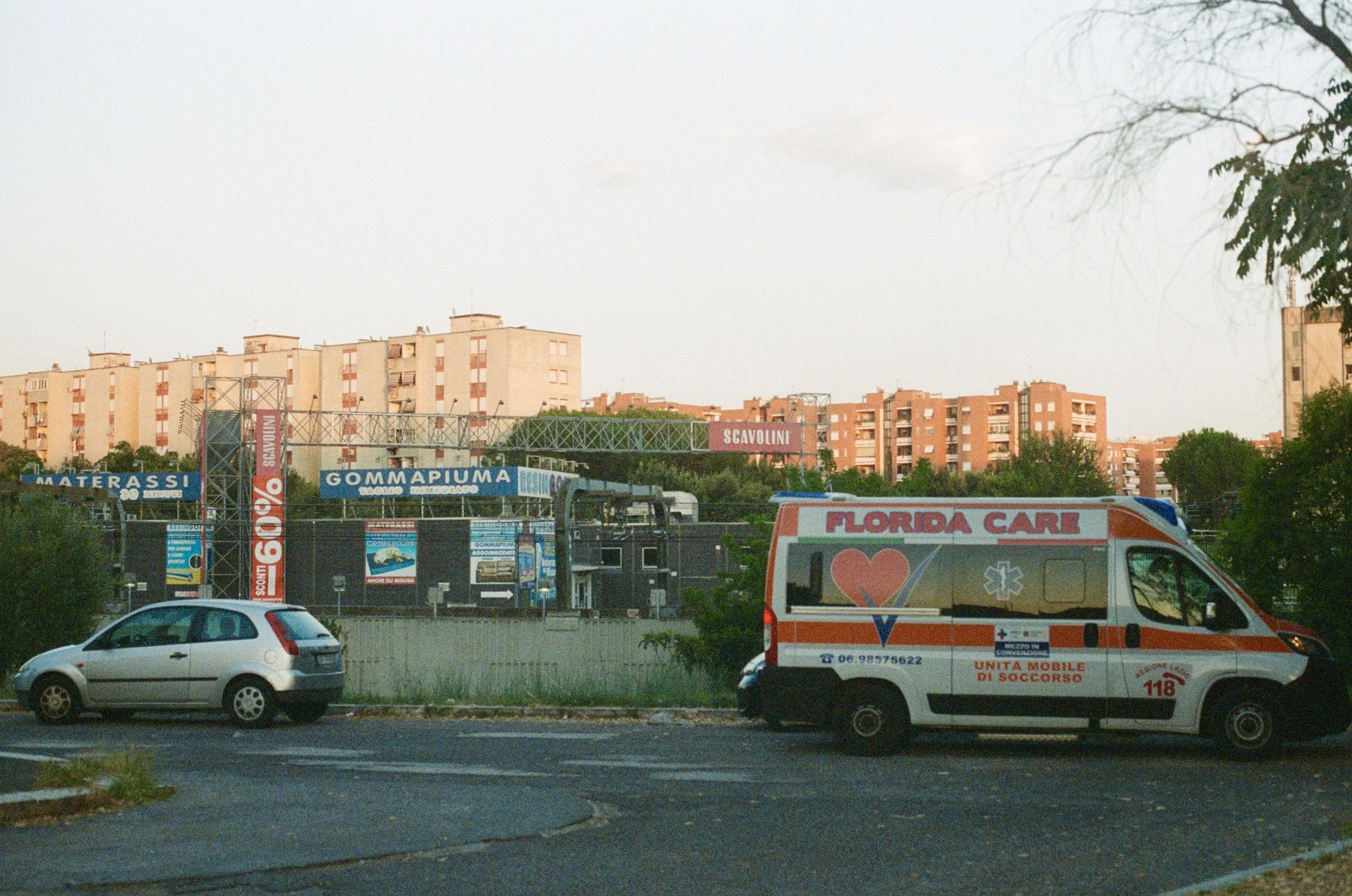 A white car and an ambulance labeled 'Florida Care' parked on a street in front of a shopping center with signs for water and rubber products, and residential apartment buildings in the background during daylight.