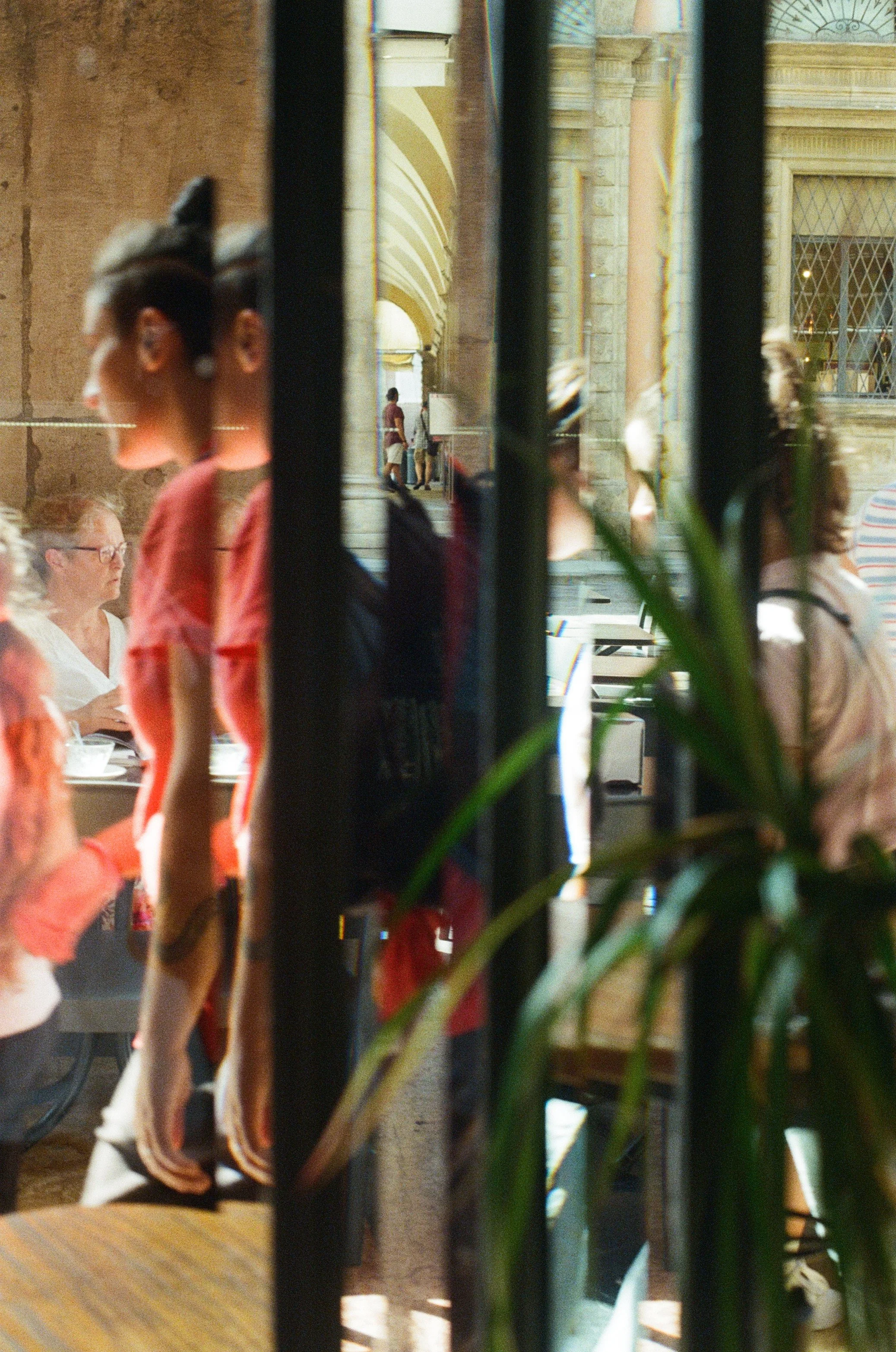 View through a window or glass partition showing waitstaff in red shirts serving diners in a busy restaurant with ornate architecture and large windows.