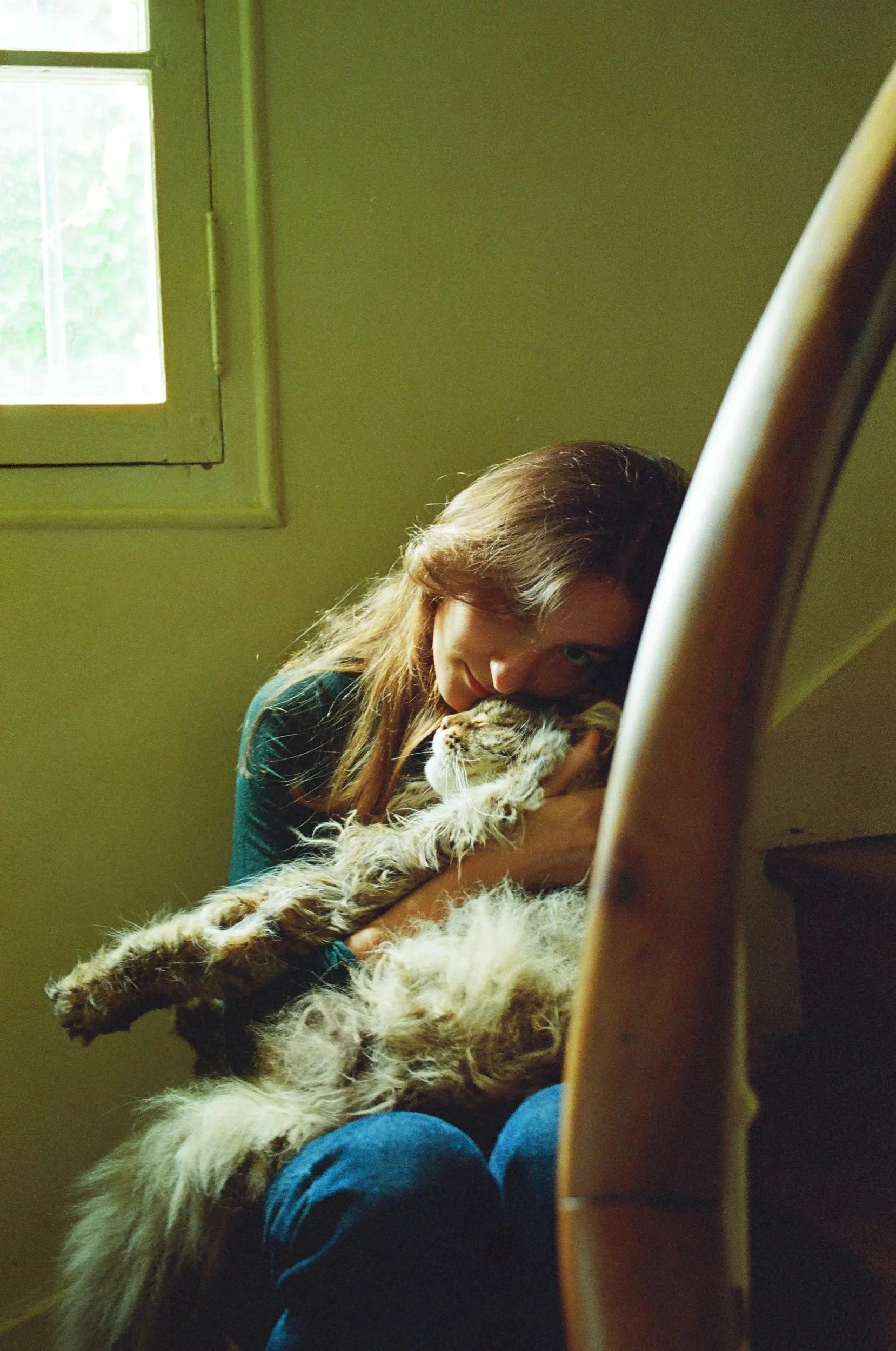A woman sitting near a window, holding and cuddling an adorable, fluffy cat on her lap.