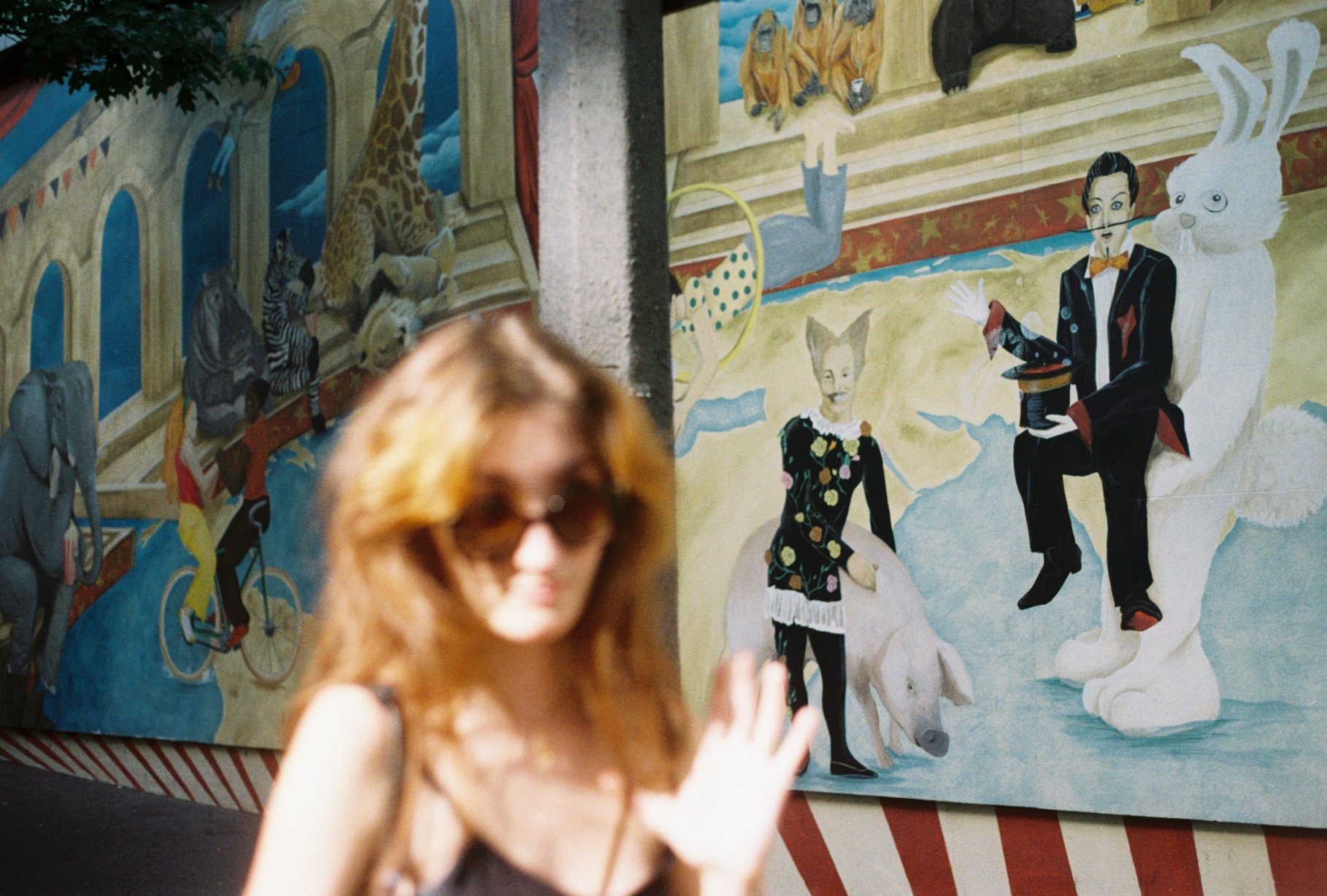 A woman with red hair wearing sunglasses in front of a colorful circus-themed mural with animals, clowns, and a magician.