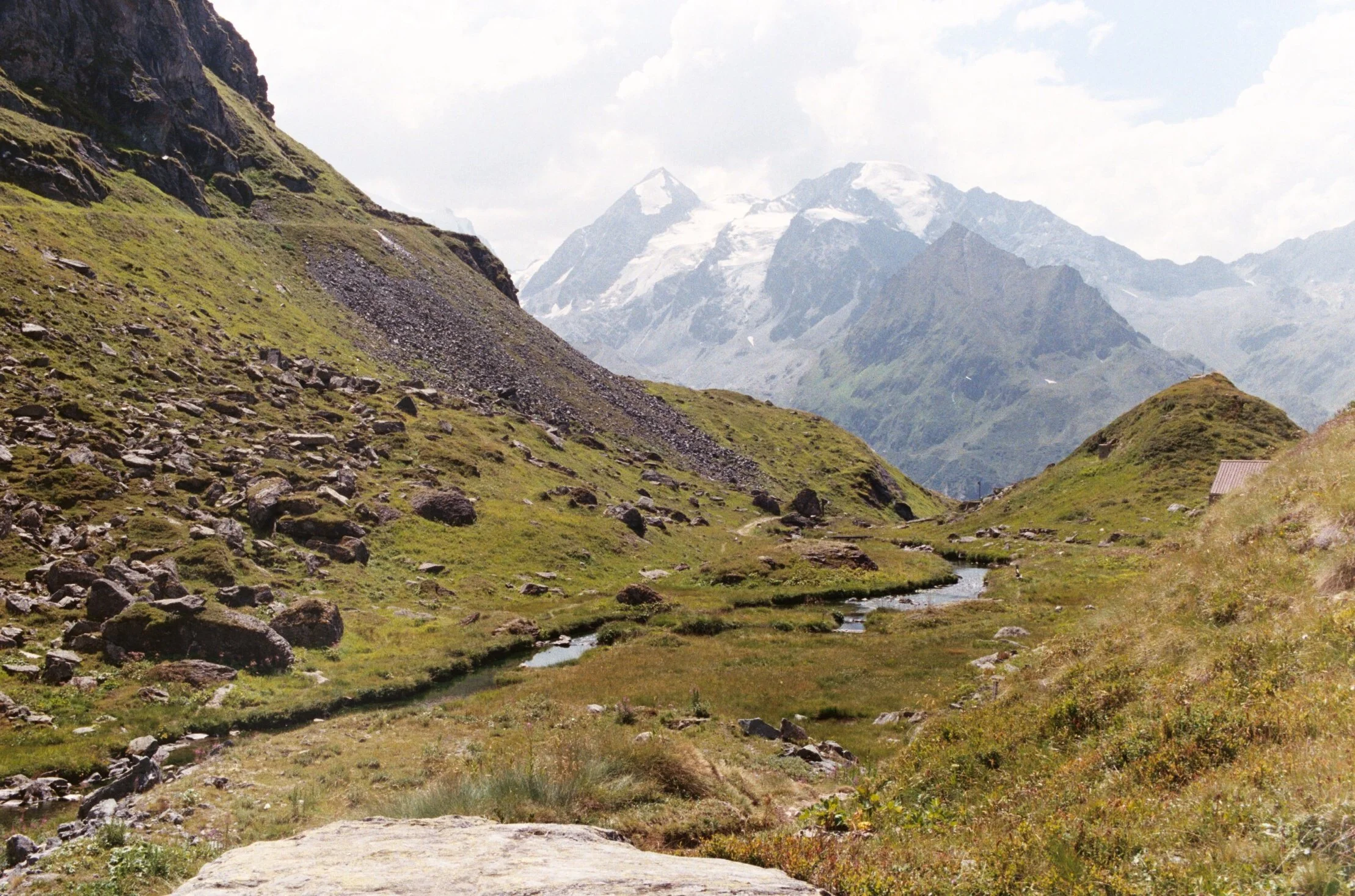 mountain landscape with snow-capped peaks, green hills, rocks, and a small stream.