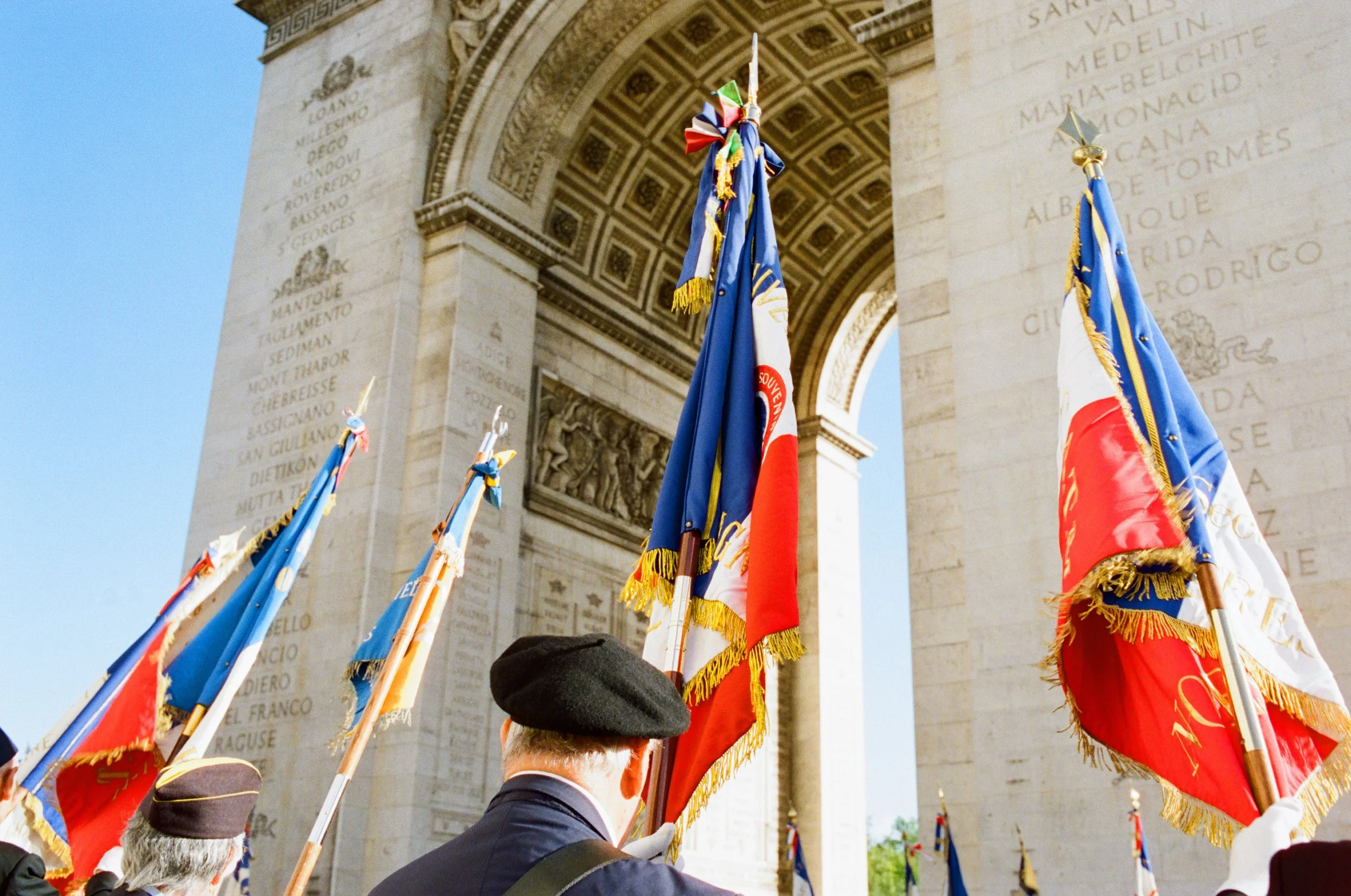 People holding flags in front of the Arc de Triomphe in Paris, France, in a commemorative or patriotic event.