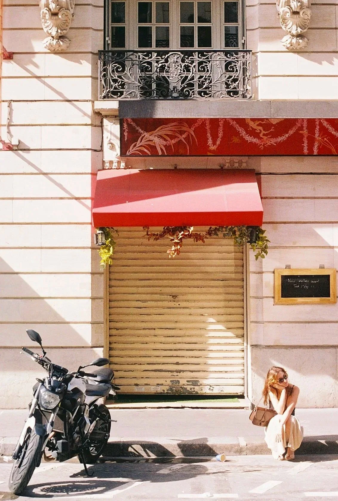 A woman sitting on the sidewalk beside a parked motorcycle, with a closed storefront with a red awning and a blackboard sign on the wall in the background.