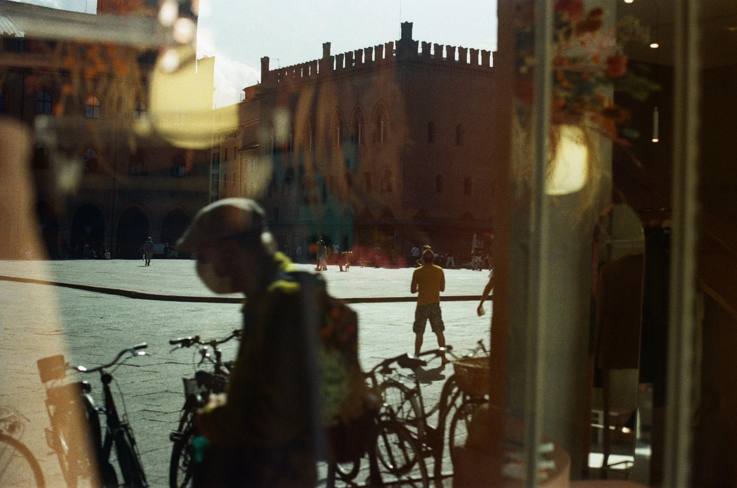 A reflection of a city square with a historic brick building and people walking, seen through a glass window that also shows bicycles and a person wearing a face mask inside.