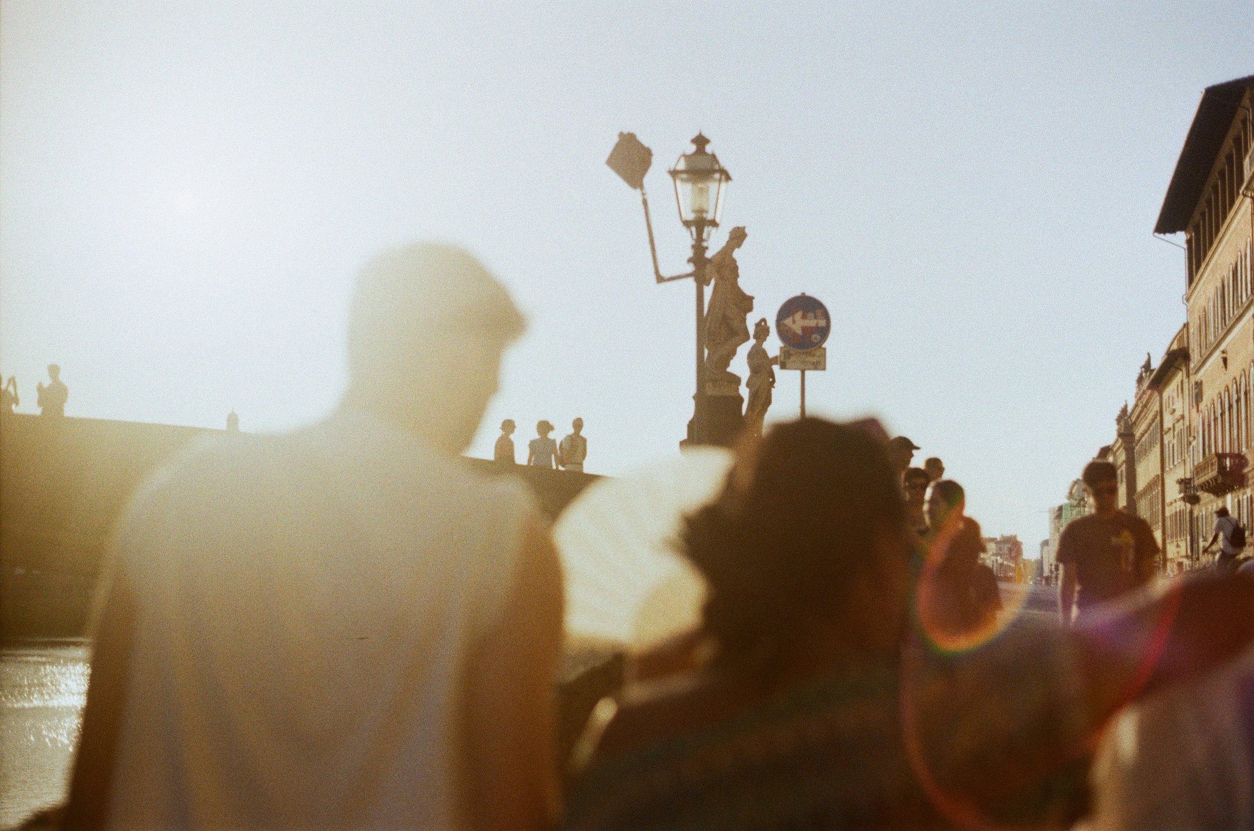 Crowd of people walking along a street during sunset, with silhouettes of buildings and a decorative street lamp with statues in the background.