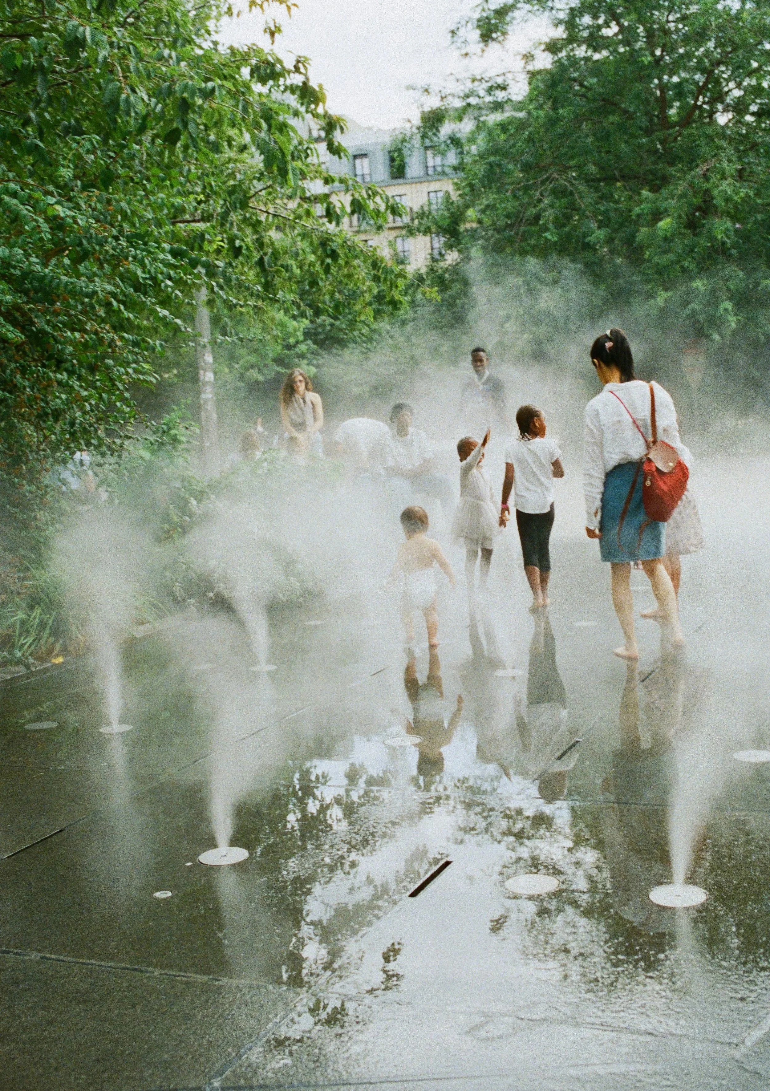 People, including children and adults, enjoying a foggy water fountain in a park, with trees and buildings in the background.