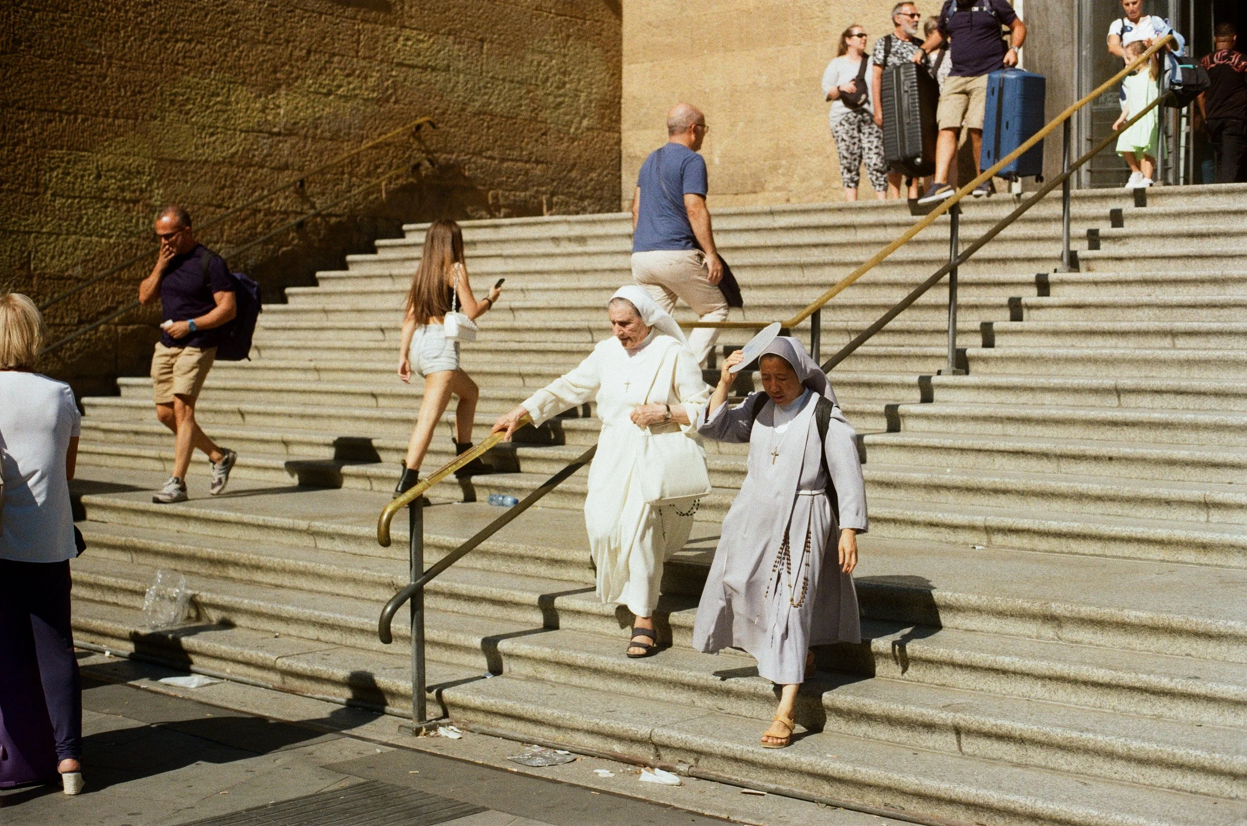 Two nuns descending city steps, one shielding eyes with hand, with tourists around, some carrying luggage.