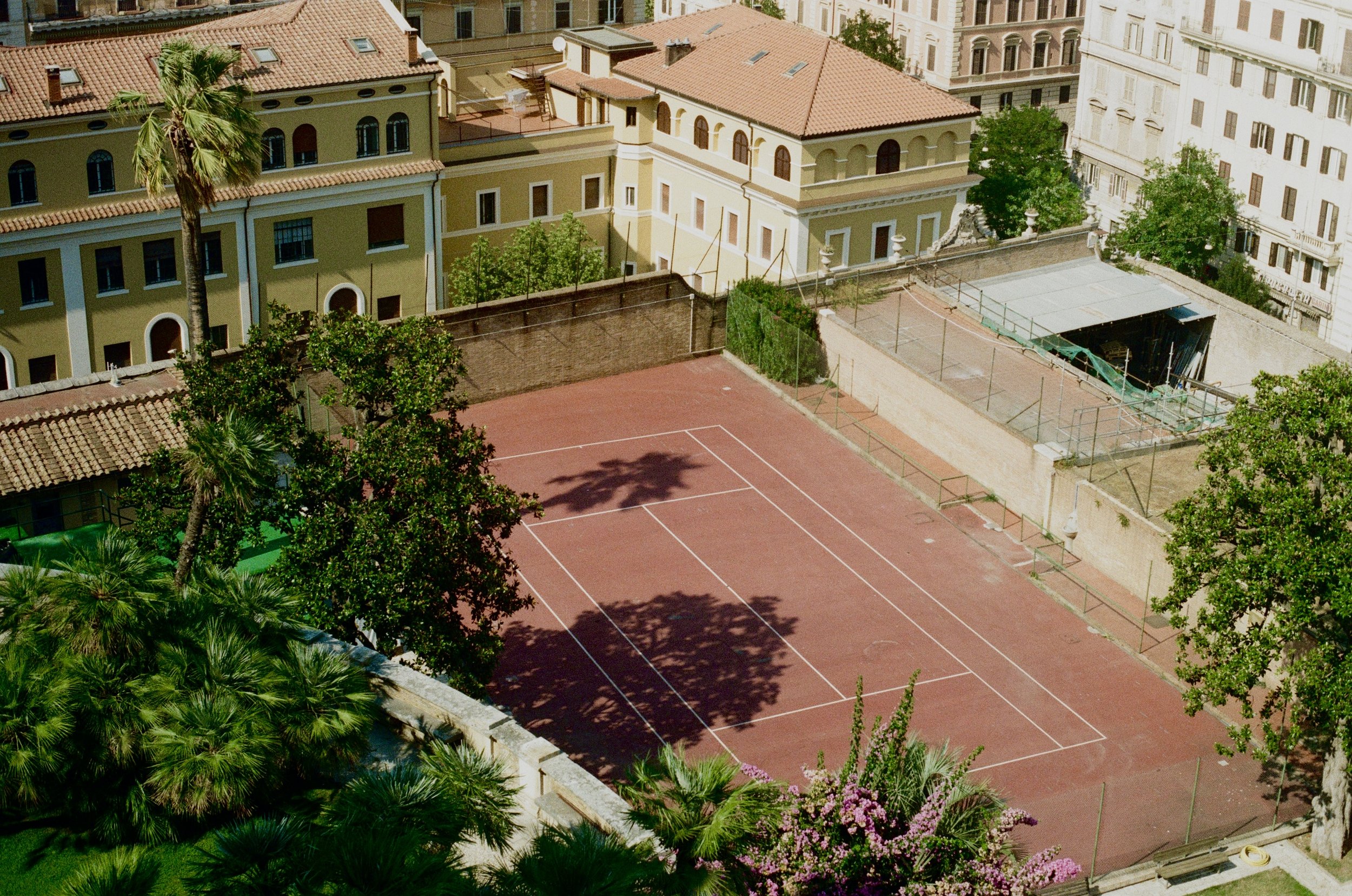 A small empty red clay tennis court surrounded by trees and nearby buildings with tiled roofs and windows.