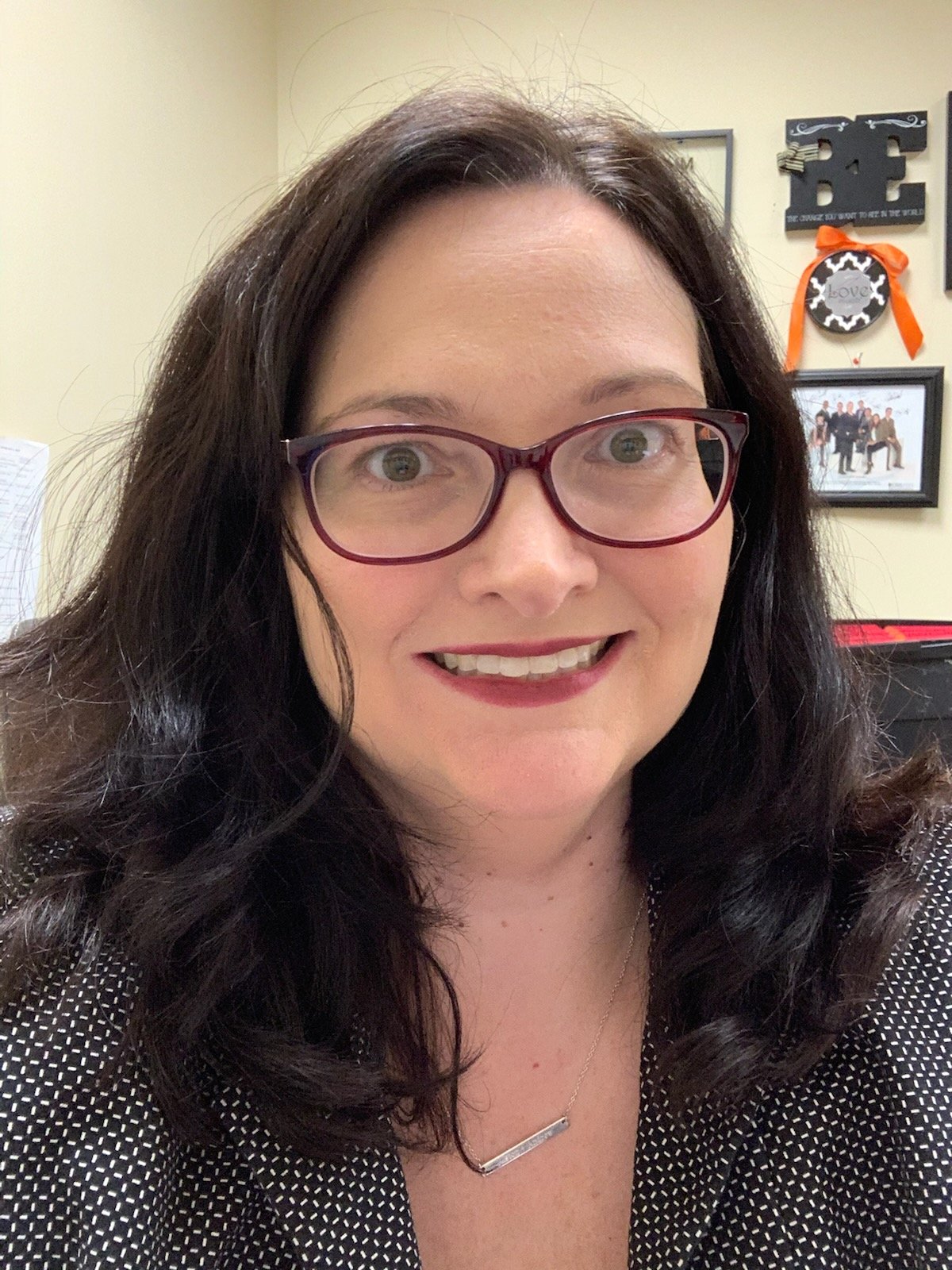A woman with dark brown hair and glasses smiling in an indoor office setting.