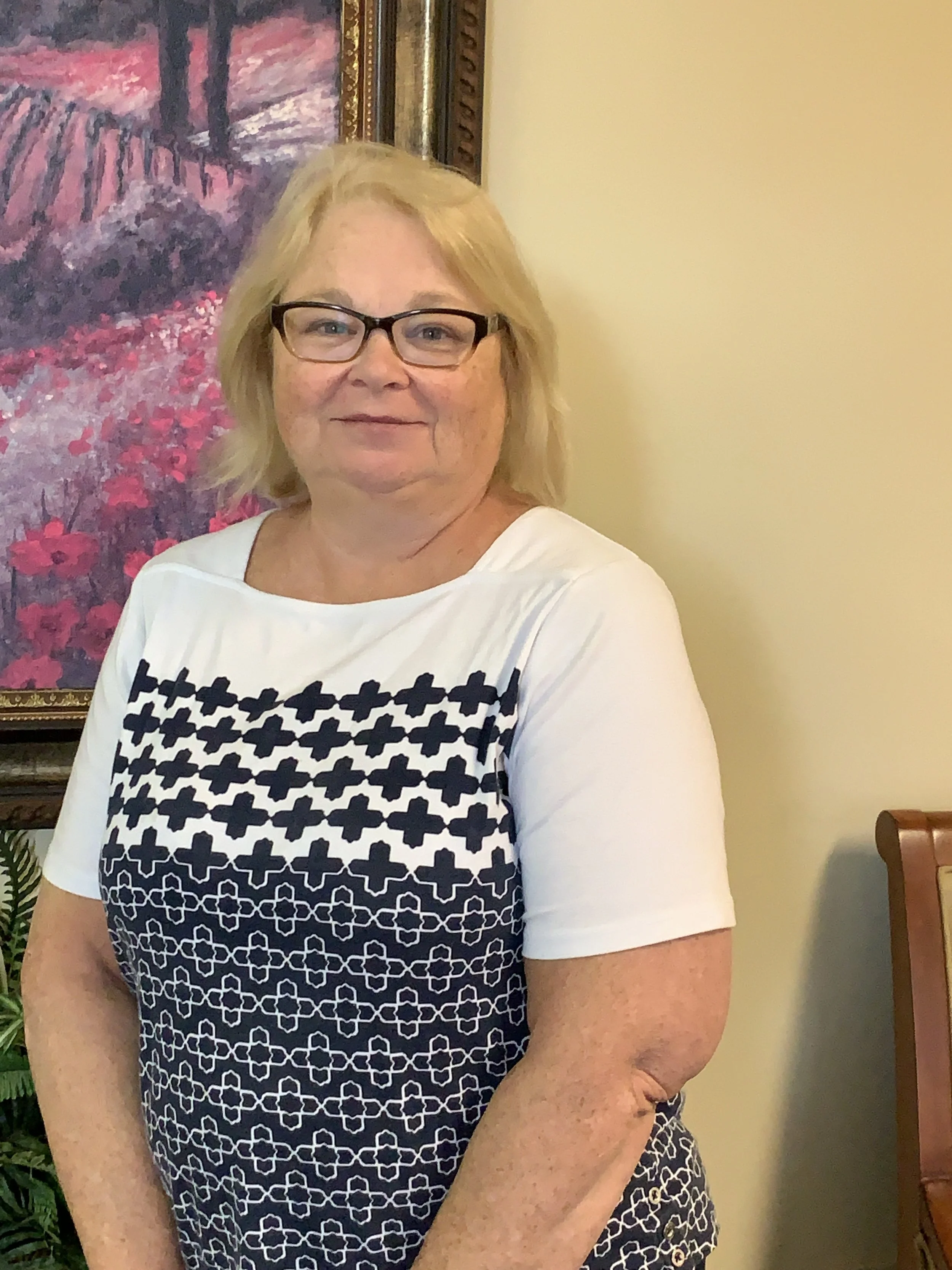 A middle-aged woman with blonde hair, glasses, and light skin standing indoors. She is wearing a white and navy patterned top and smiling at the camera. In the background, there is a framed painting of a landscape with pink and purple flowers and greenery.