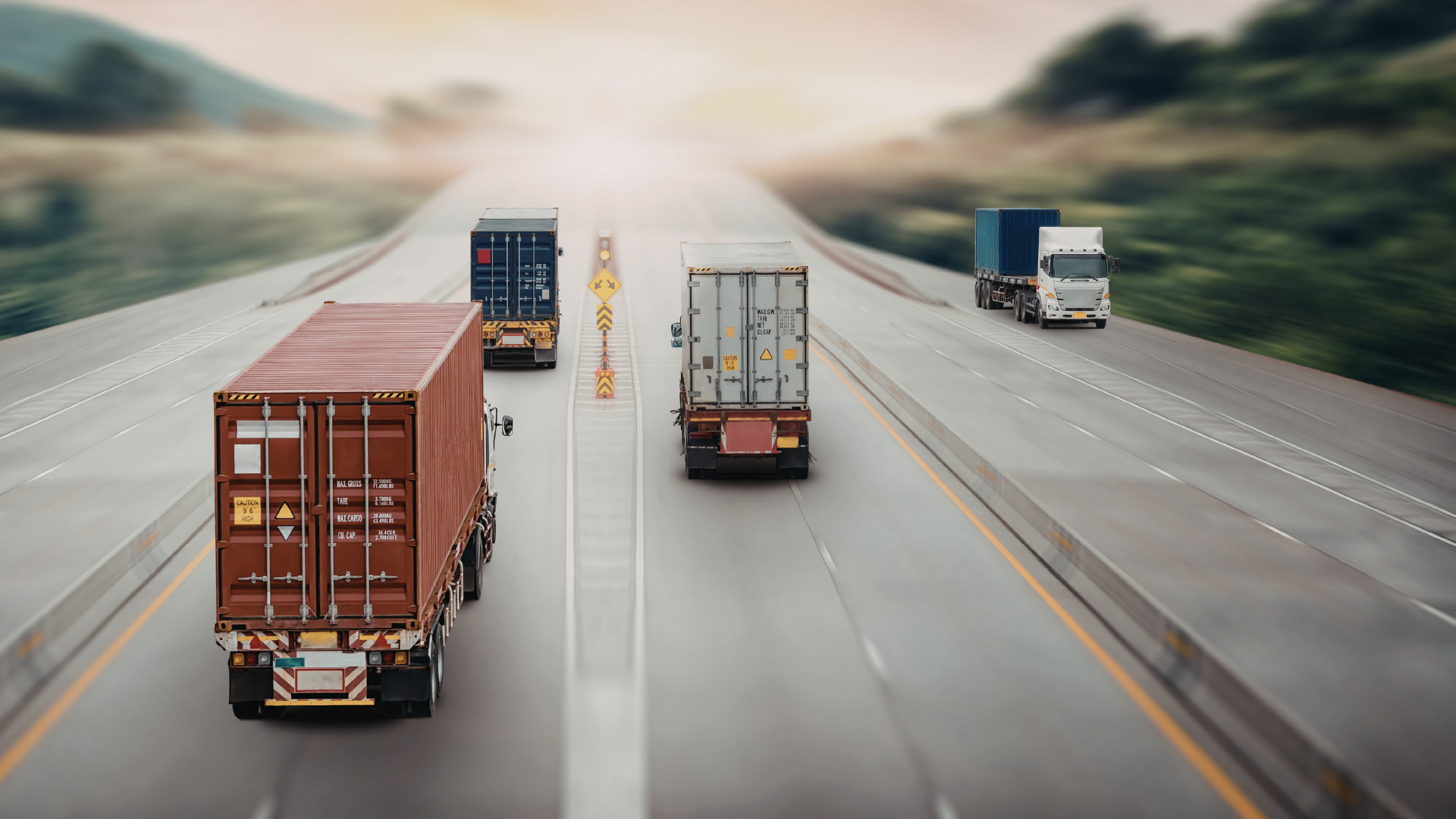 Several large trucks carrying shipping containers driving on a highway during sunset or sunrise.