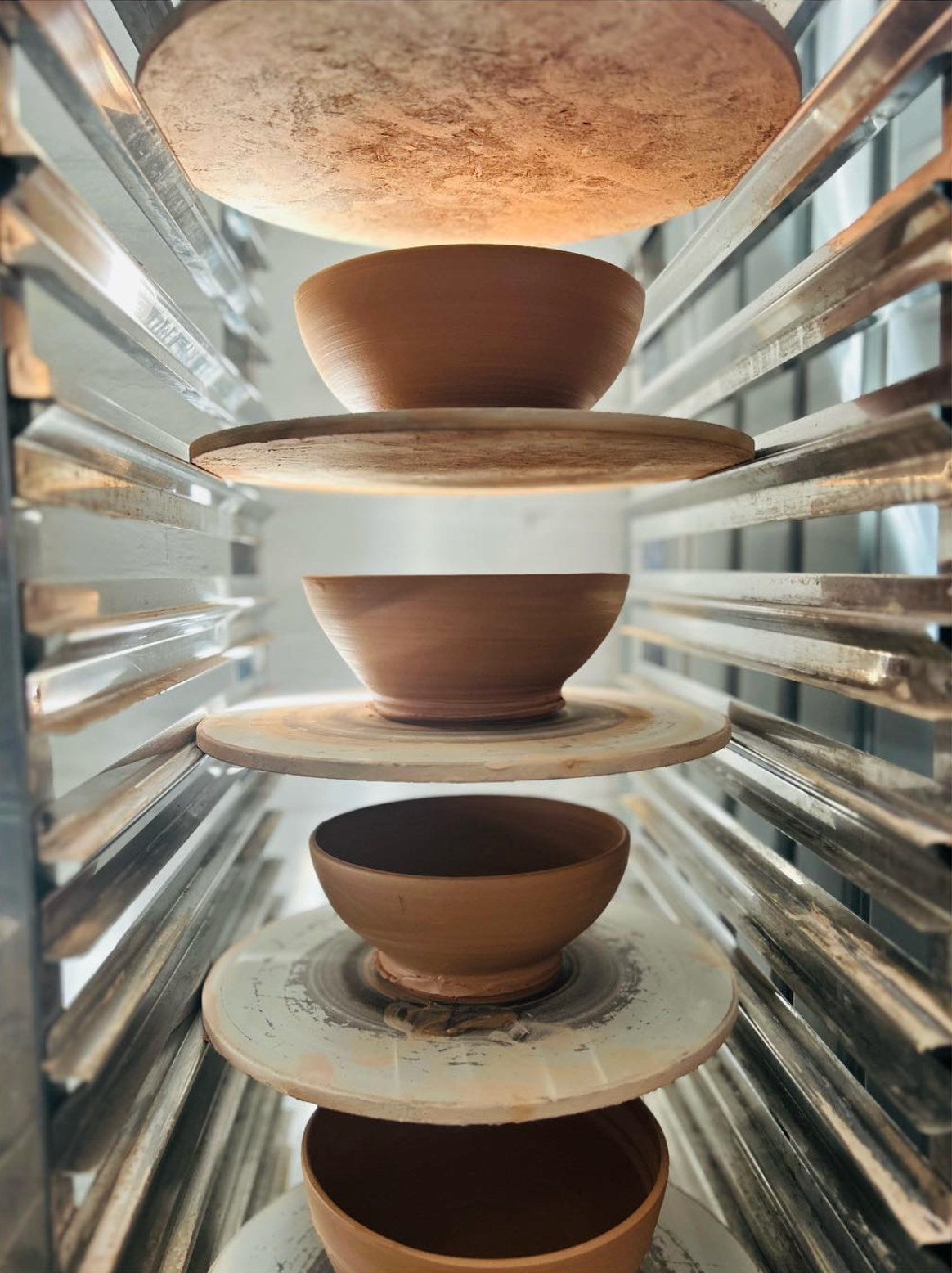 Multiple clay bowls in various stages of drying on a rotating rack in a pottery kiln. The bowls are on circular platforms between metal supports.