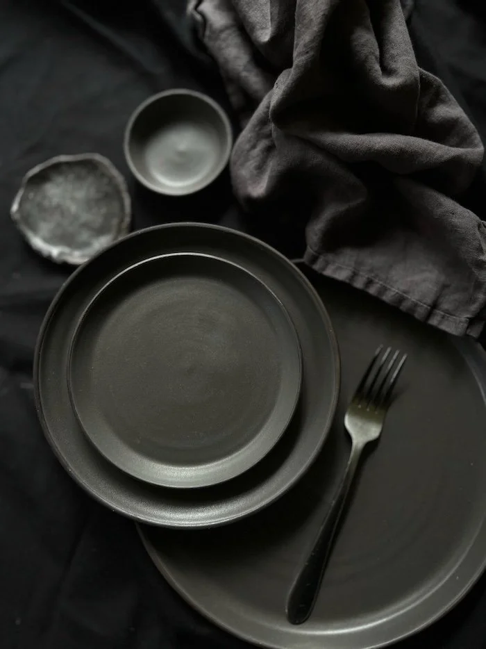 Flat lay of black ceramic dinnerware including a large plate, a smaller plate, and a bowl, with a black fork on the large plate, alongside a gray cloth napkin, a small gray bowl, and a small dish, all on a black surface.