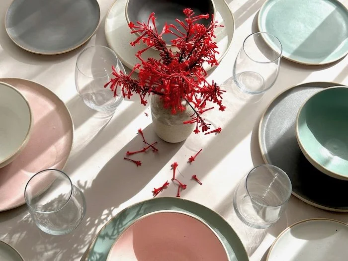 A dining table set with pastel-colored plates and clear glasses, with a white vase holding bright red decorative branches in the center, and some of the branches scattered on the table.