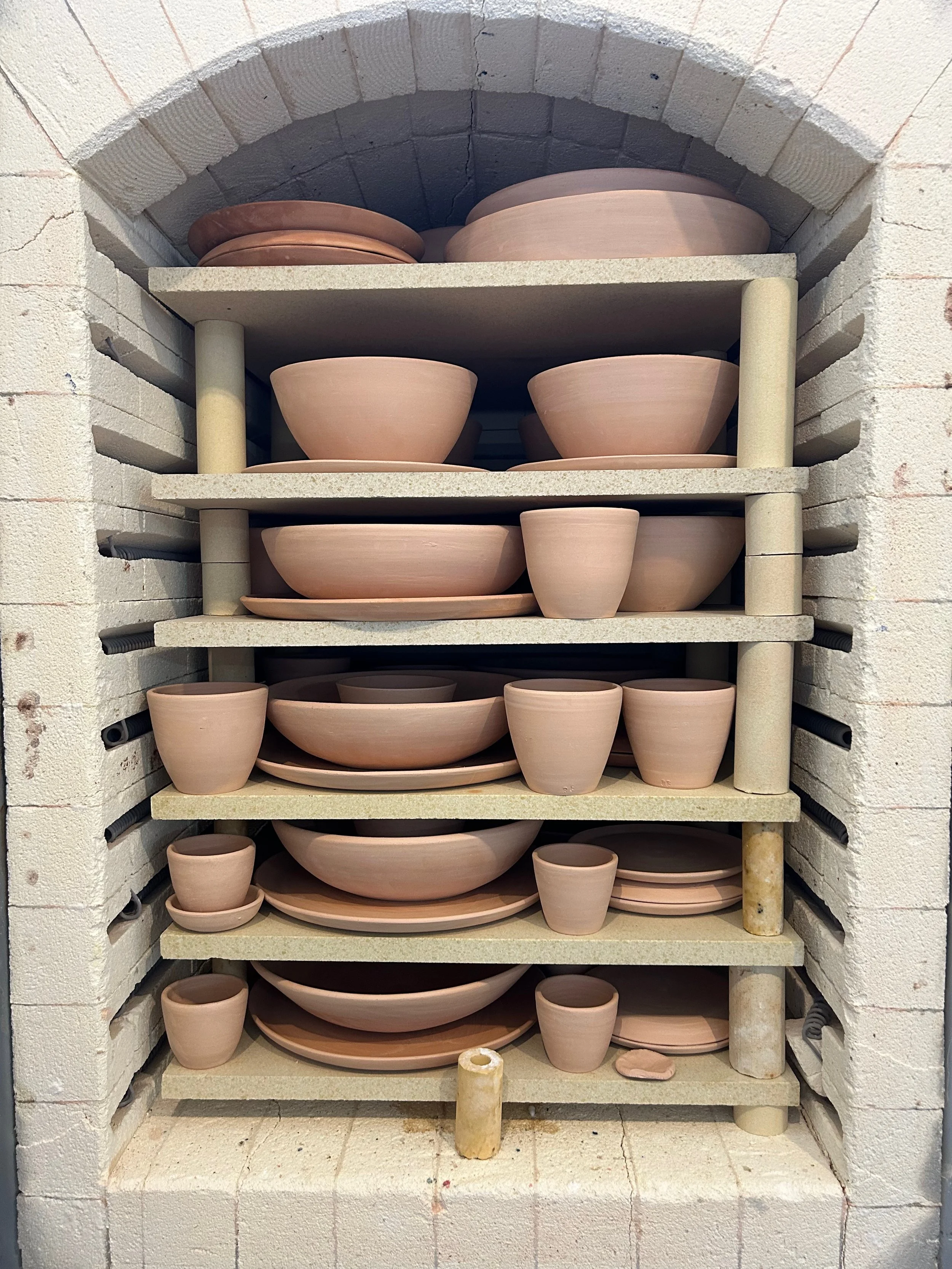 Shelves filled with unglazed pottery bowls and plates in a kiln or oven. The shelves are supported by vertical poles and positioned inside a brick structure.