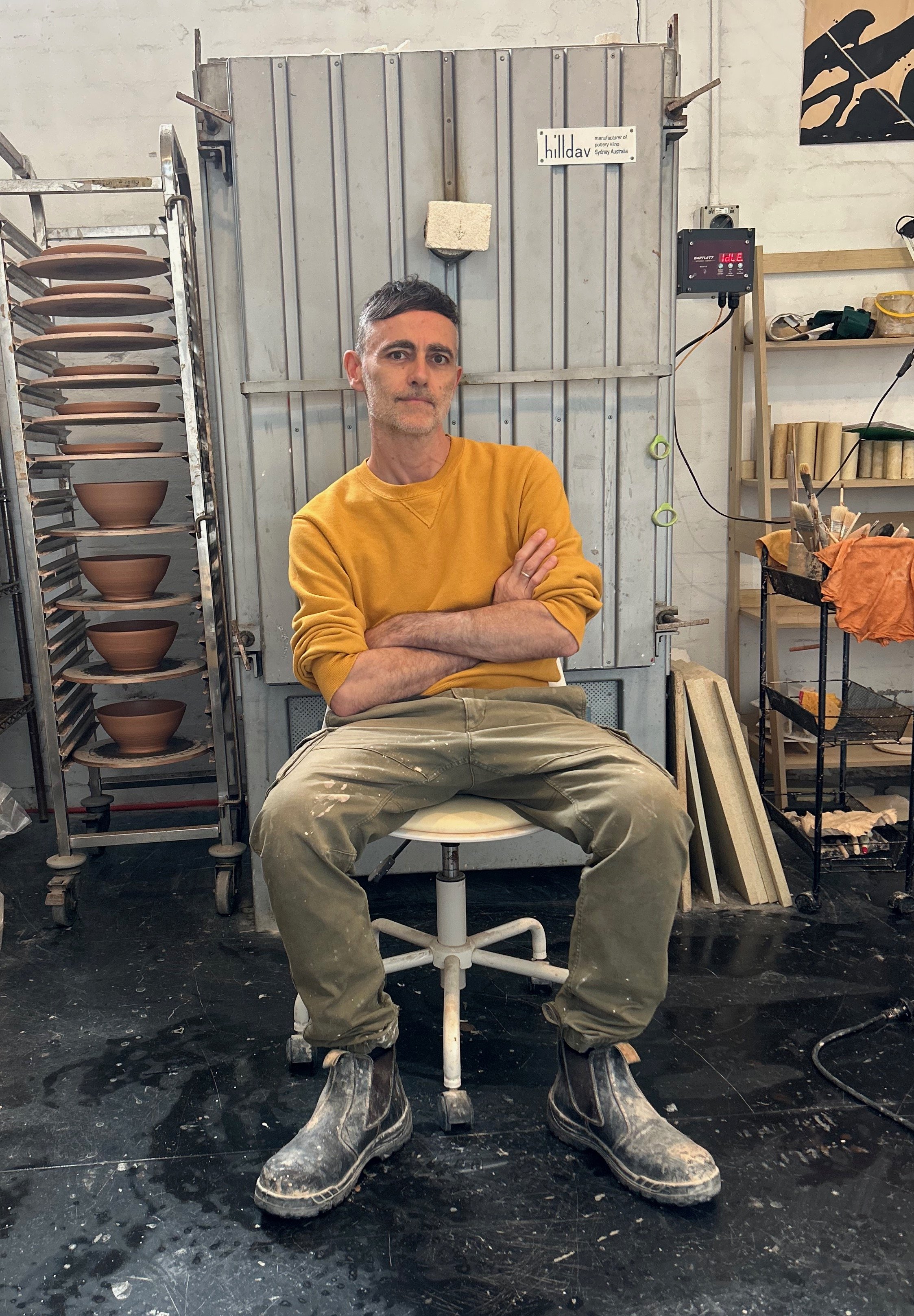 A man in a yellow sweatshirt and green pants sits with arms crossed in a pottery studio, surrounded by shelves of unfinished ceramics, tools, and equipment.