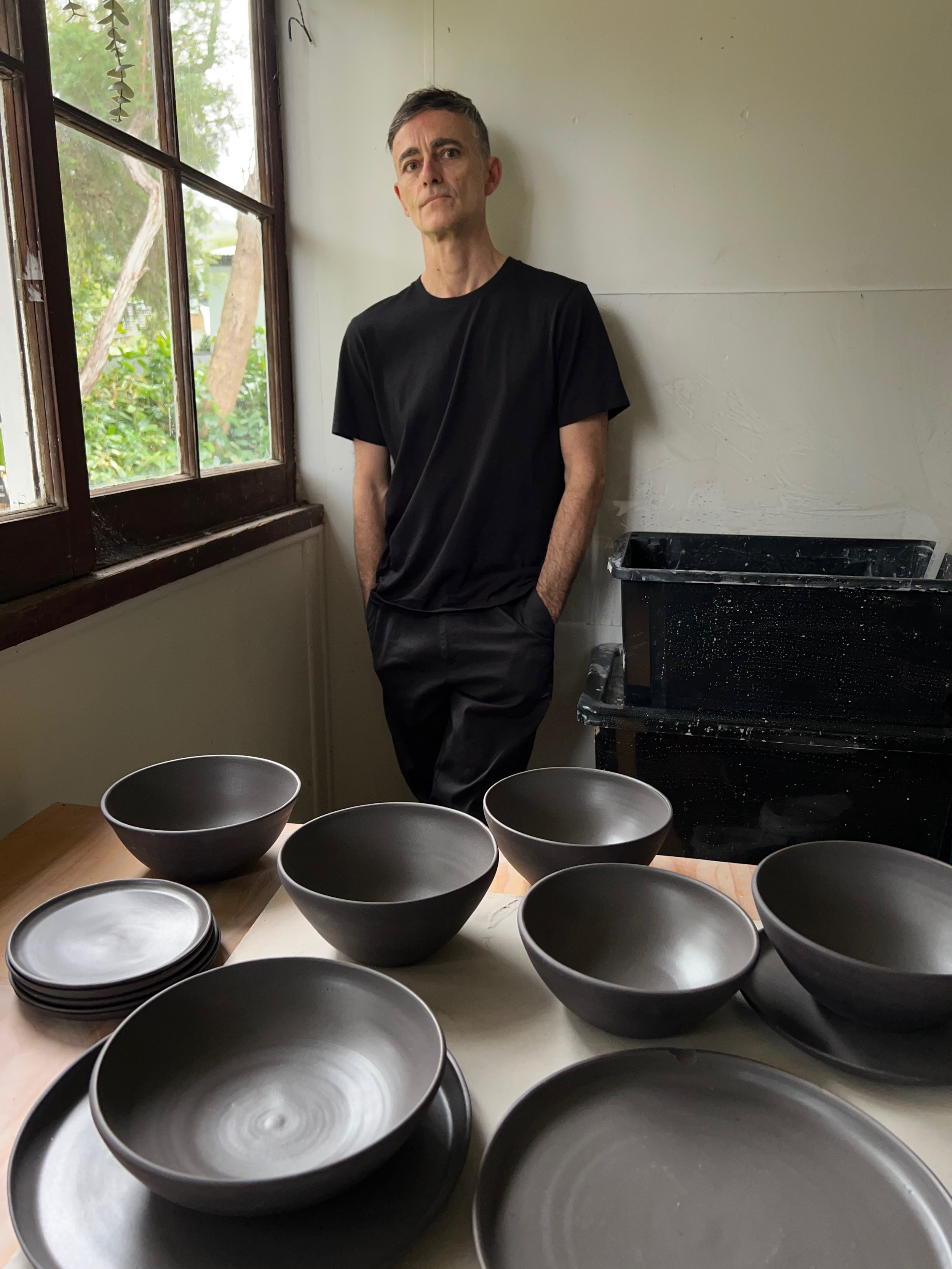 A man standing indoors near a window with green trees outside. In the foreground, there are several black bowls of various sizes on a table.