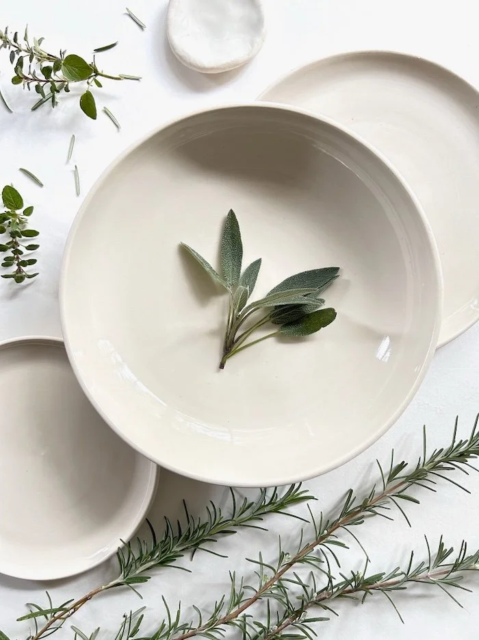 Cream-colored ceramic bowls with green herbs and sprigs of rosemary on a white surface.