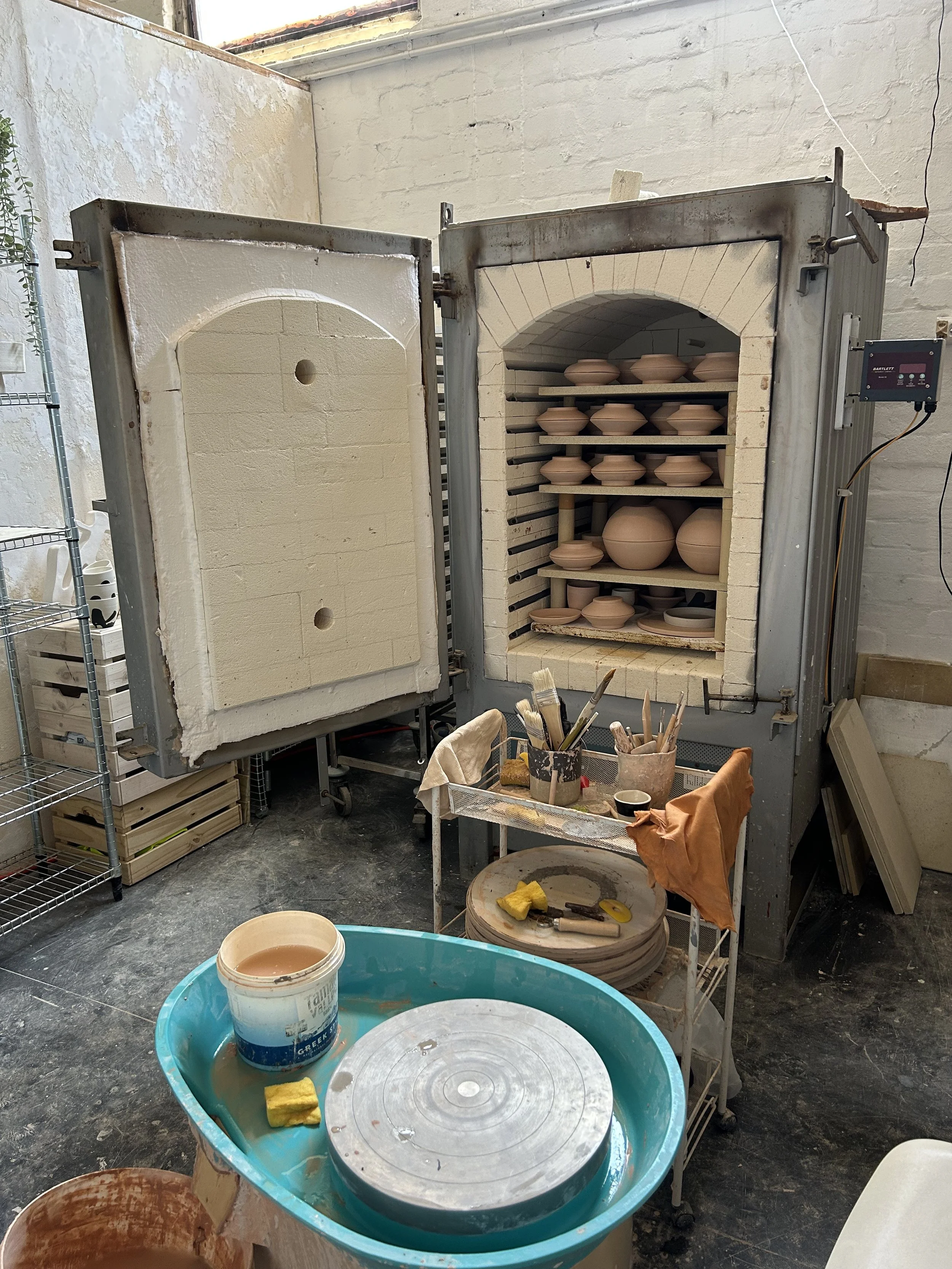 A pottery kiln in a studio with various unglazed clay bowls and pots inside on shelves. There are tools and brushes on a small table in front. The studio has a concrete floor and unfinished walls.