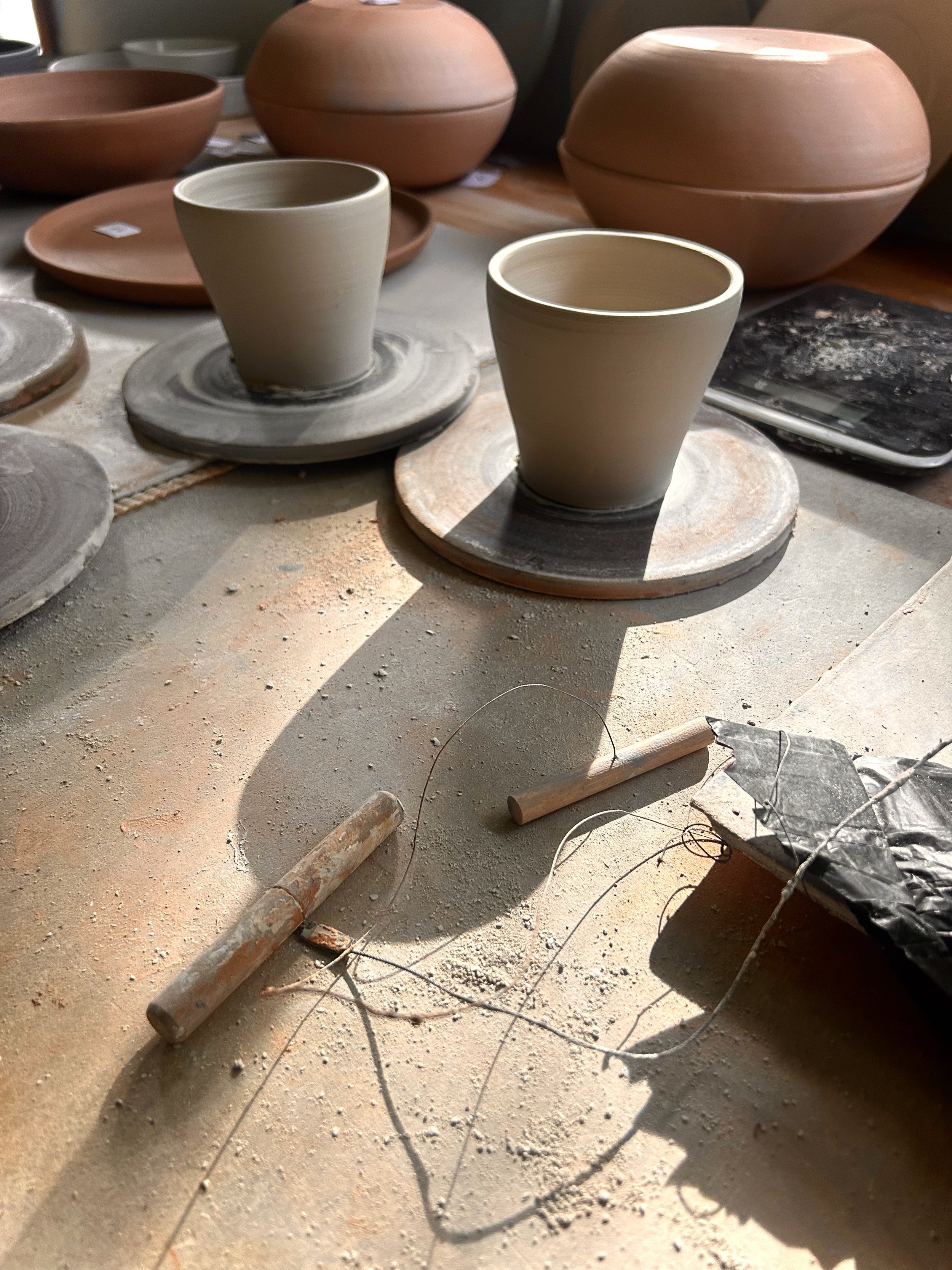 A cluttered workspace with pottery pieces, including two cups on pottery wheels, and various tools, wires, and pottery in progress on a dusty surface.
