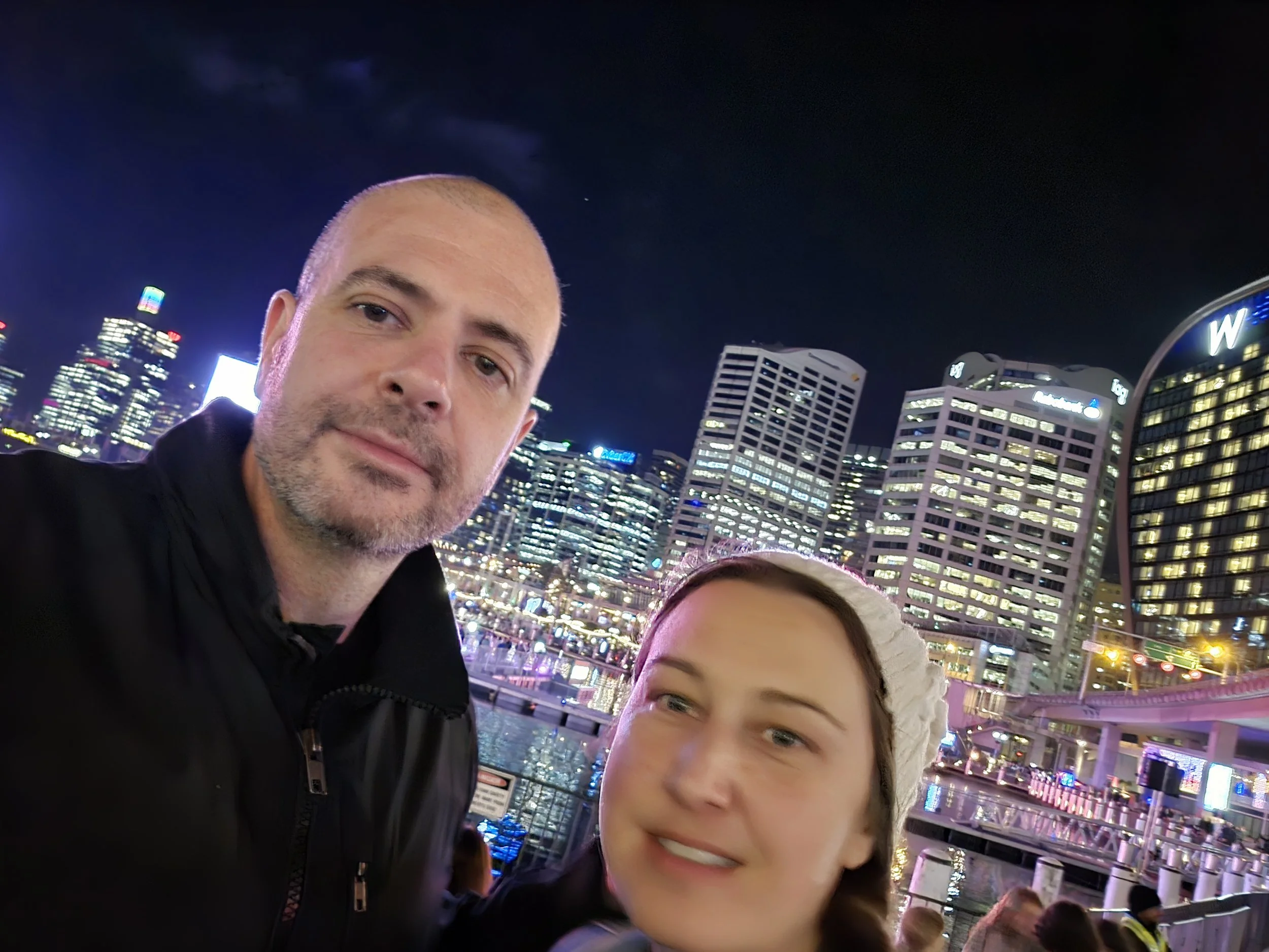 A man and young girl taking a selfie at night in a city with illuminated skyscrapers, water, and a curved bridge.