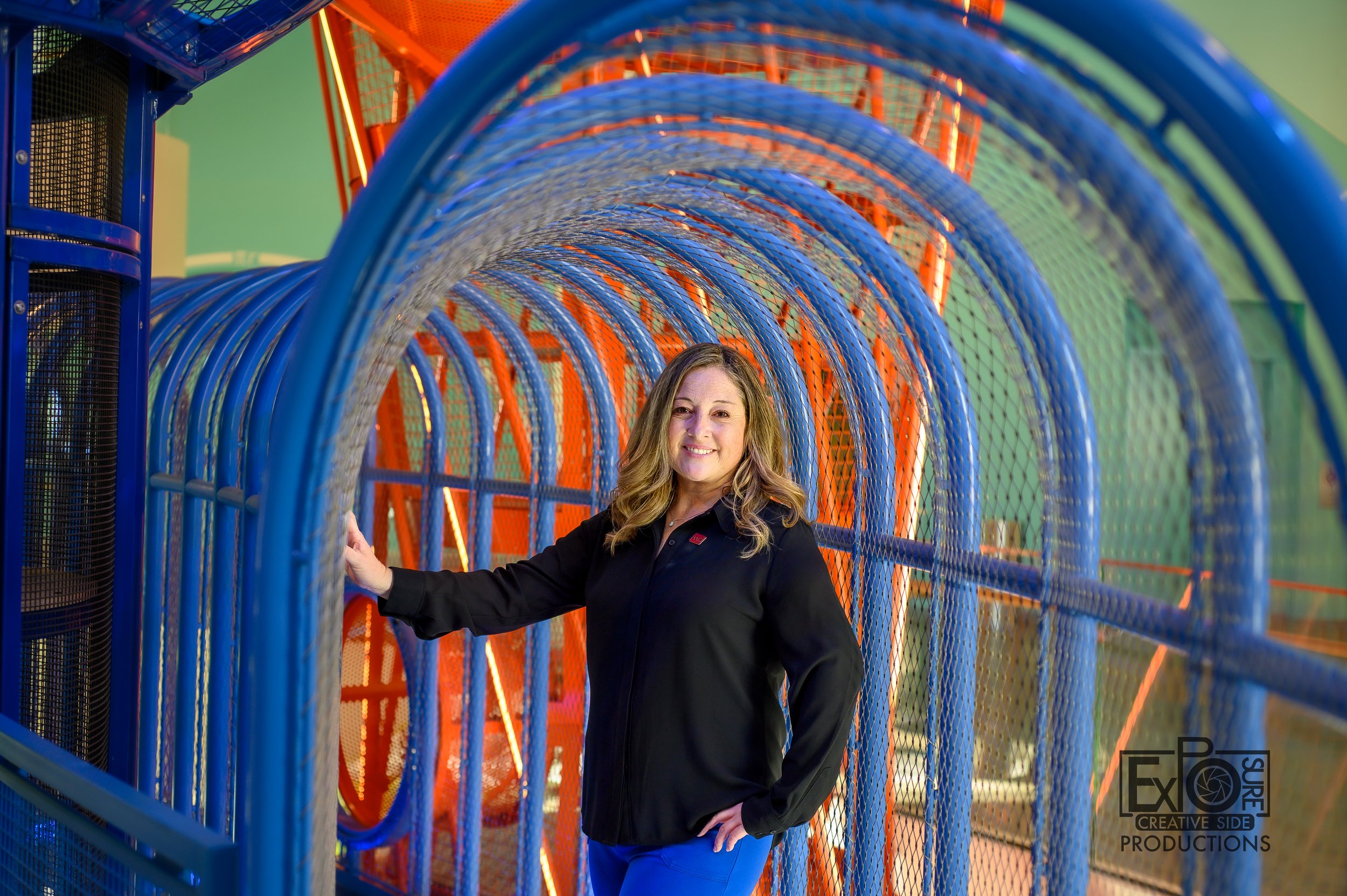 A woman standing inside a colorful indoor playground tunnel structure, smiling and holding onto the blue bars, with orange and blue colors surrounding her.