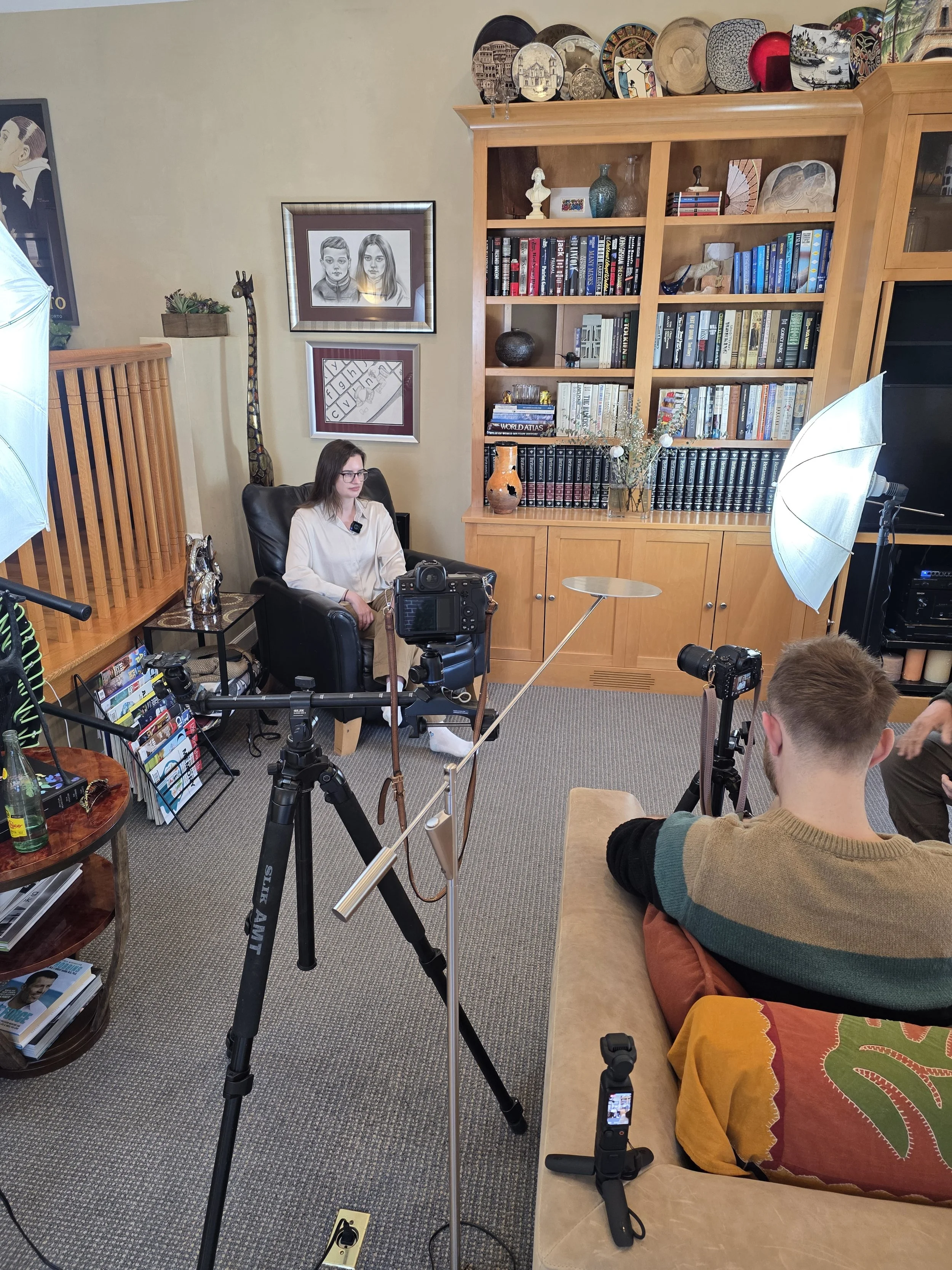 A woman in a white sweater sits in a black armchair in a room with bookshelves, framed artwork, and decorative items. There are cameras, lighting equipment, and crew members preparing for an interview or video recording.