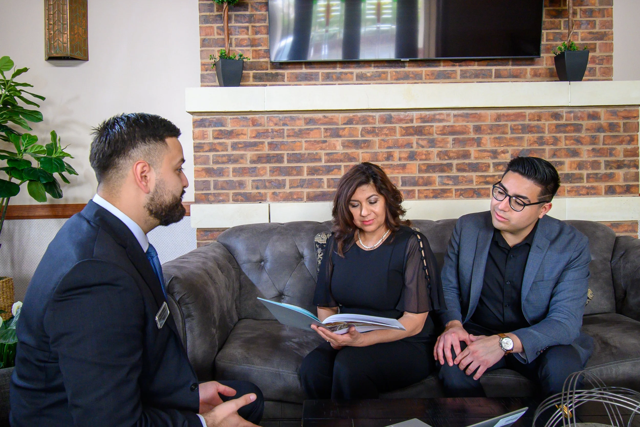 Three people sitting on a brown leather couch in a living room, reviewing documents. Two men in business suits and a woman in black attire are engaged in discussion. The room has a brick wall, a mounted TV, and decorative plants.