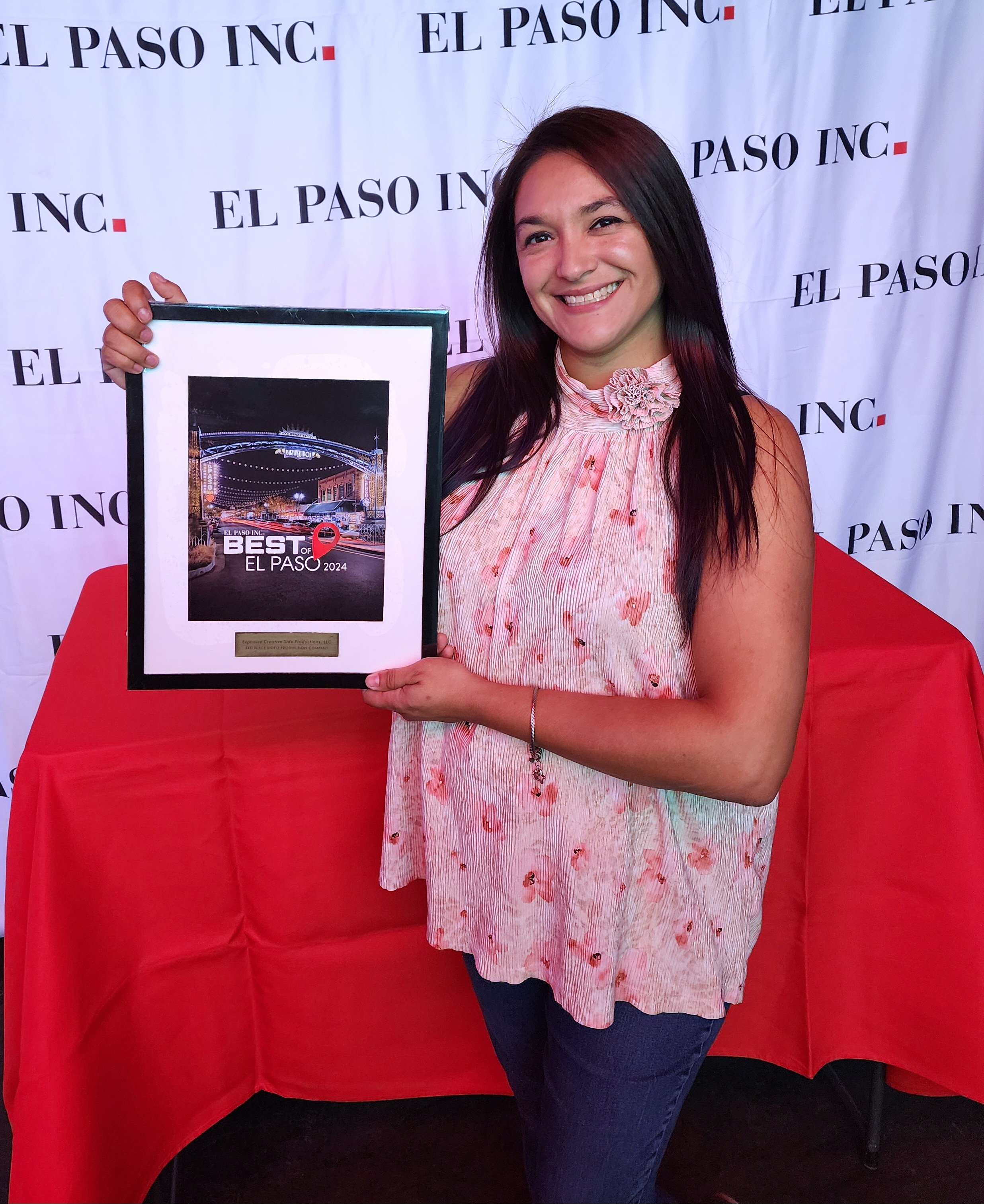A woman smiling and holding a framed award in front of a backdrop with 'EL PASO INC' written repeatedly. The award features a nighttime city scene with the text 'Best of El Paso 2024'.
