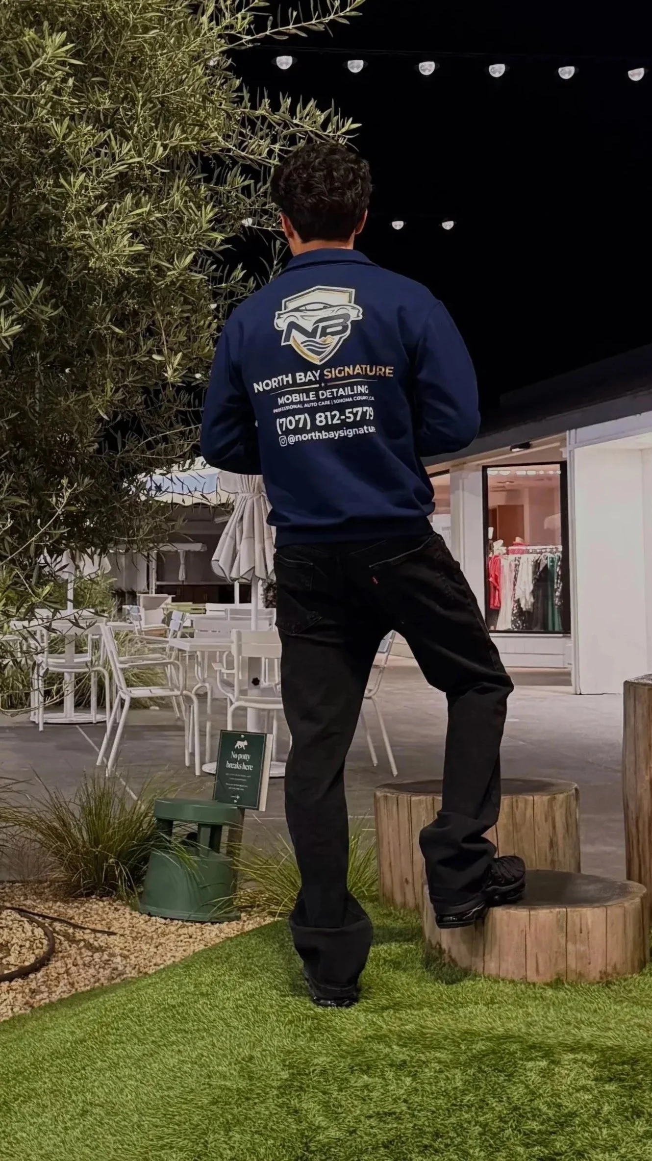 A person standing outdoors at night, wearing a dark blue jacket with business information on the back, standing on a tree stump with artificial grass and a plant nearby, and patio furniture in the background.