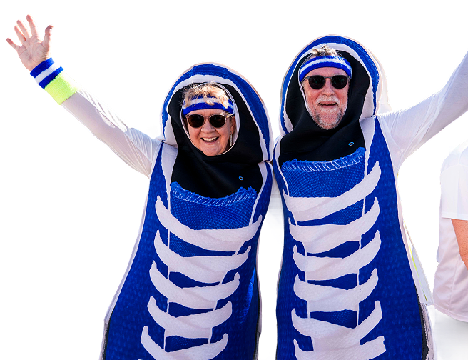 Two people dressed in skeleton costumes, wearing sunglasses and headbands, smiling with arms raised.