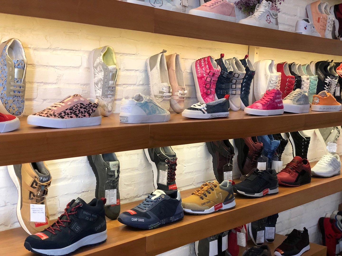 Display of various colorful sneakers on wooden shelves in a retail store, arranged in rows against a white brick wall.