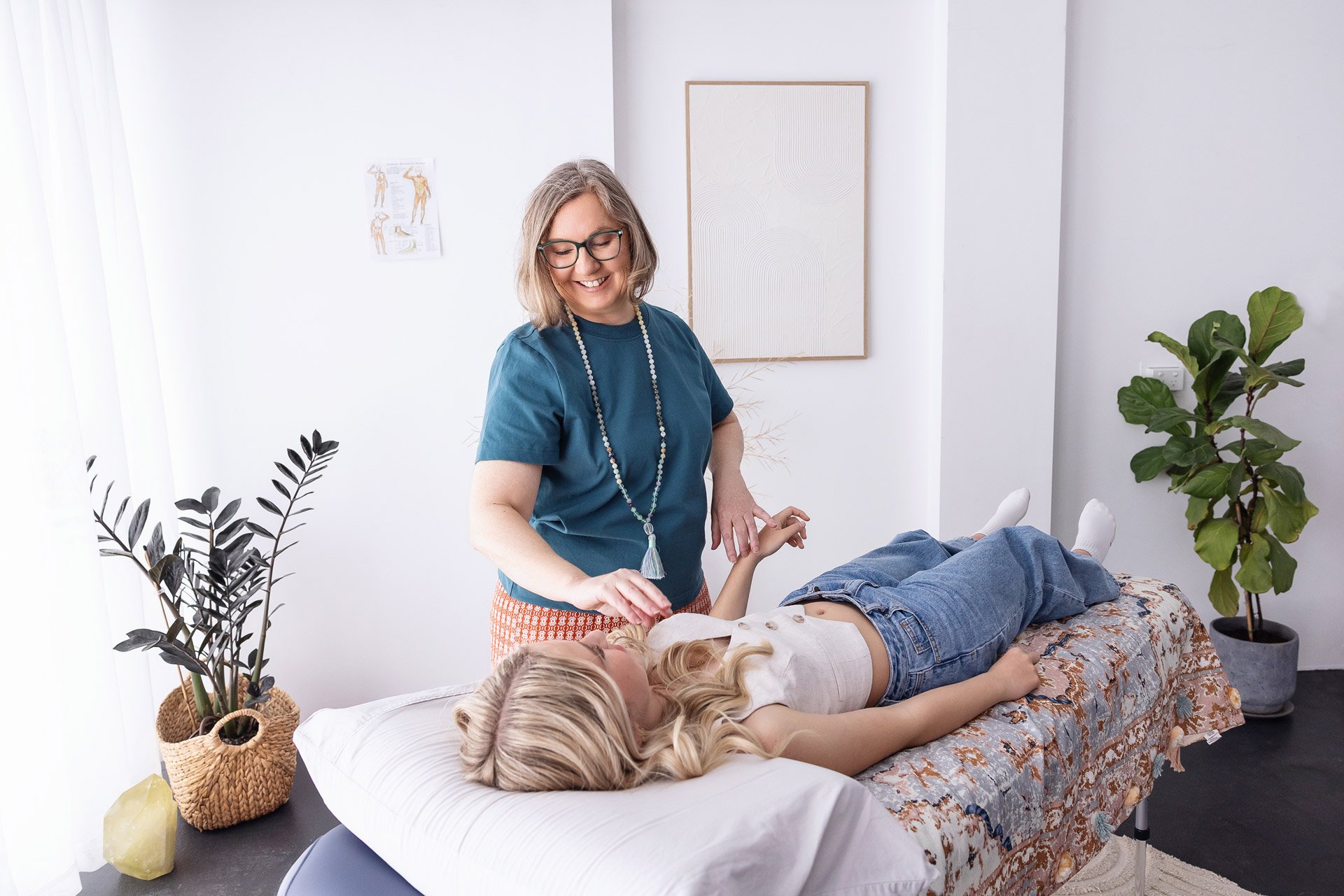 A woman receiving a therapeutic treatment or massage from a practitioner at a wellness center, with the patient lying on a treatment table surrounded by decorative plants and artwork.