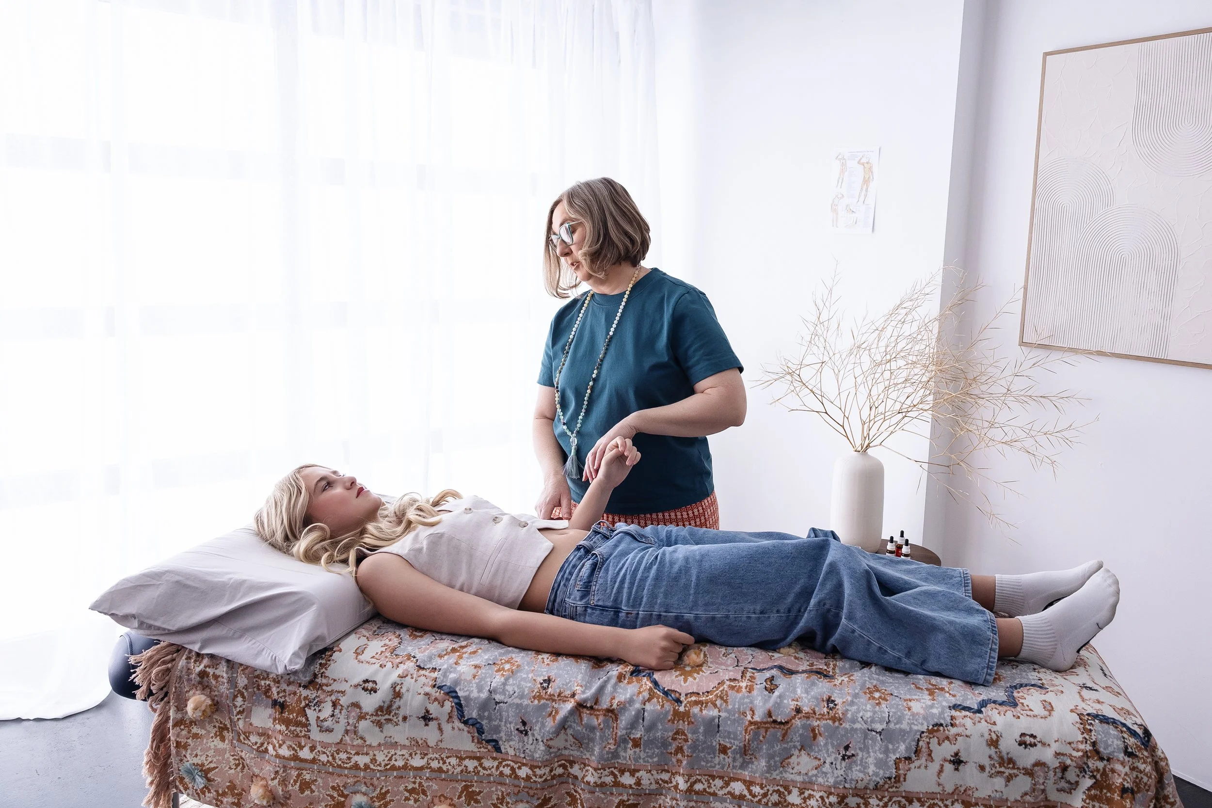 A woman lying on a massage table being massaged by another woman in a room with natural light, minimal decor, and artwork on the wall.