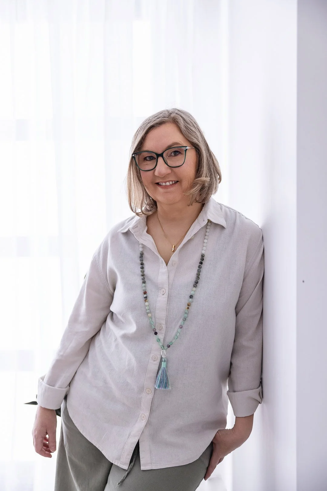 Bunbury Kinesiologist Tanya Spooner I A woman with shoulder-length gray hair, glasses, and a light-colored shirt standing indoors against a white wall and sheer curtains, smiling at the camera.