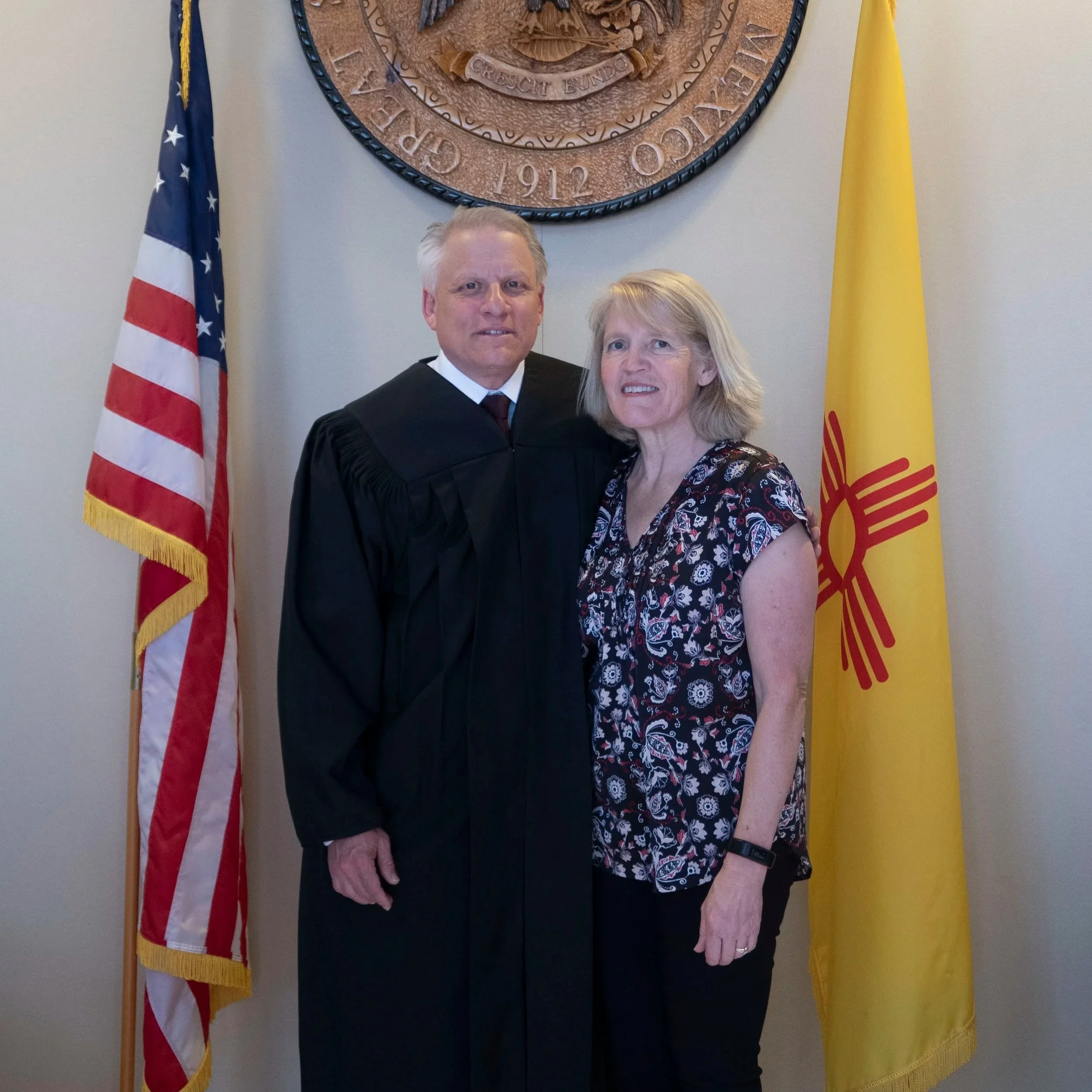 A man in a judicial robe standing next to a woman in a blouse, both standing in front of the American and New Mexico flags with a large wooden seal on the wall behind them.