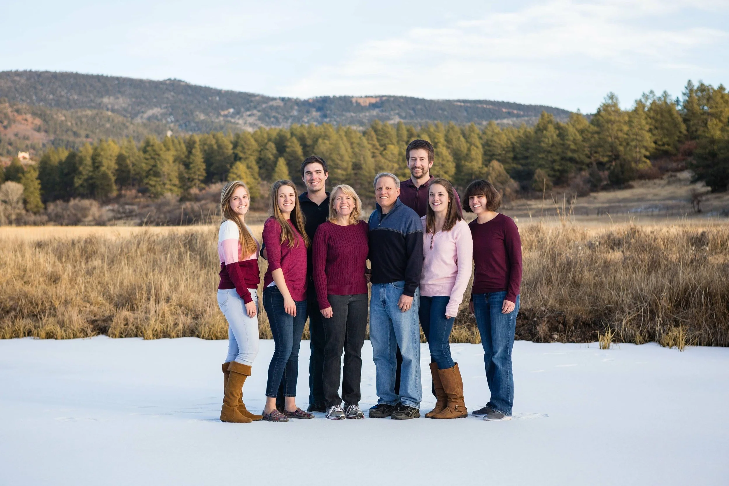 Family of eight posing outdoors on a snowy field with hills and trees in the background, all smiling and wearing winter clothing.