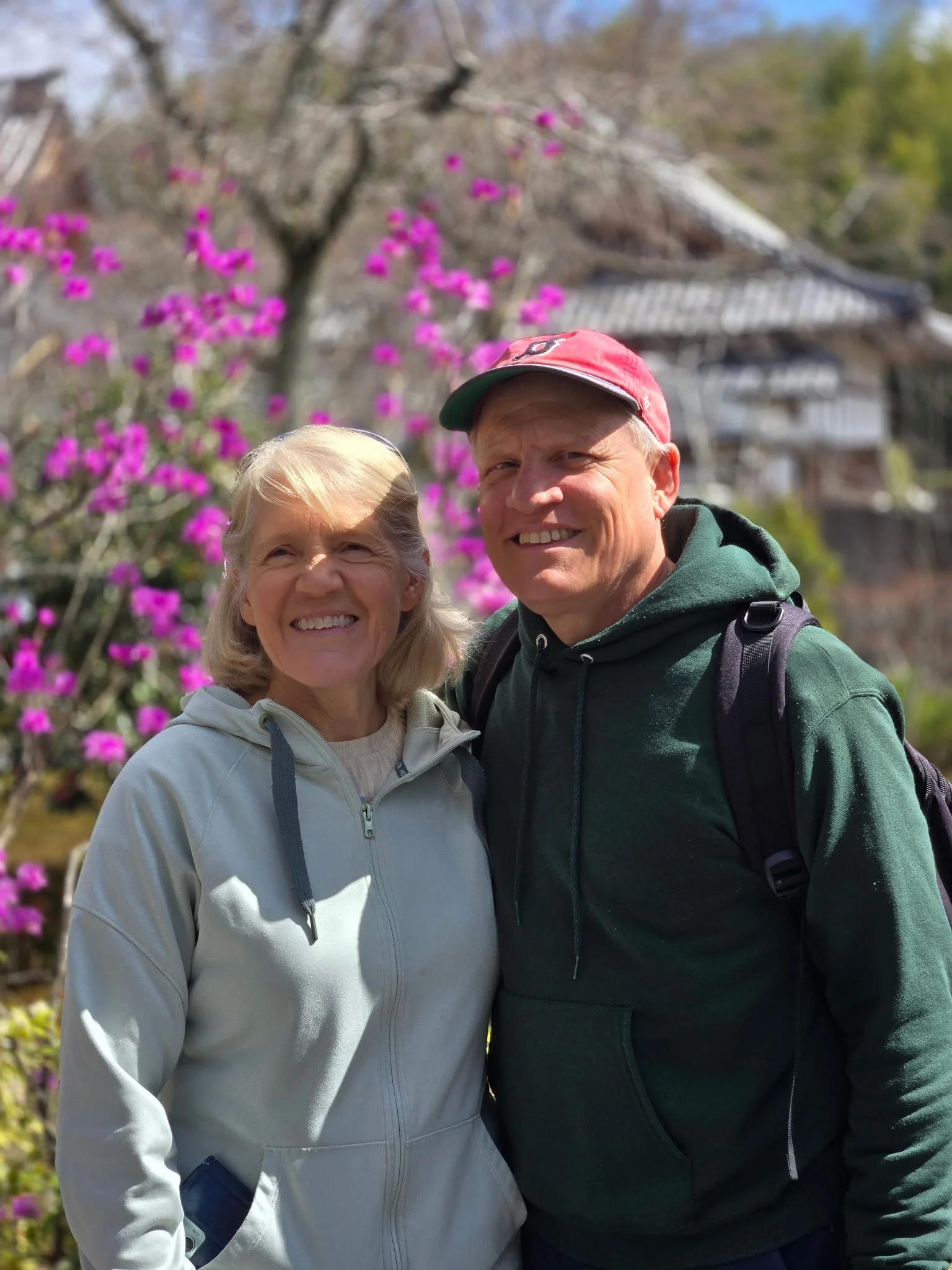 A smiling elderly woman and a smiling man stand outdoors, with a background of pink flowers, trees, and a traditional house.