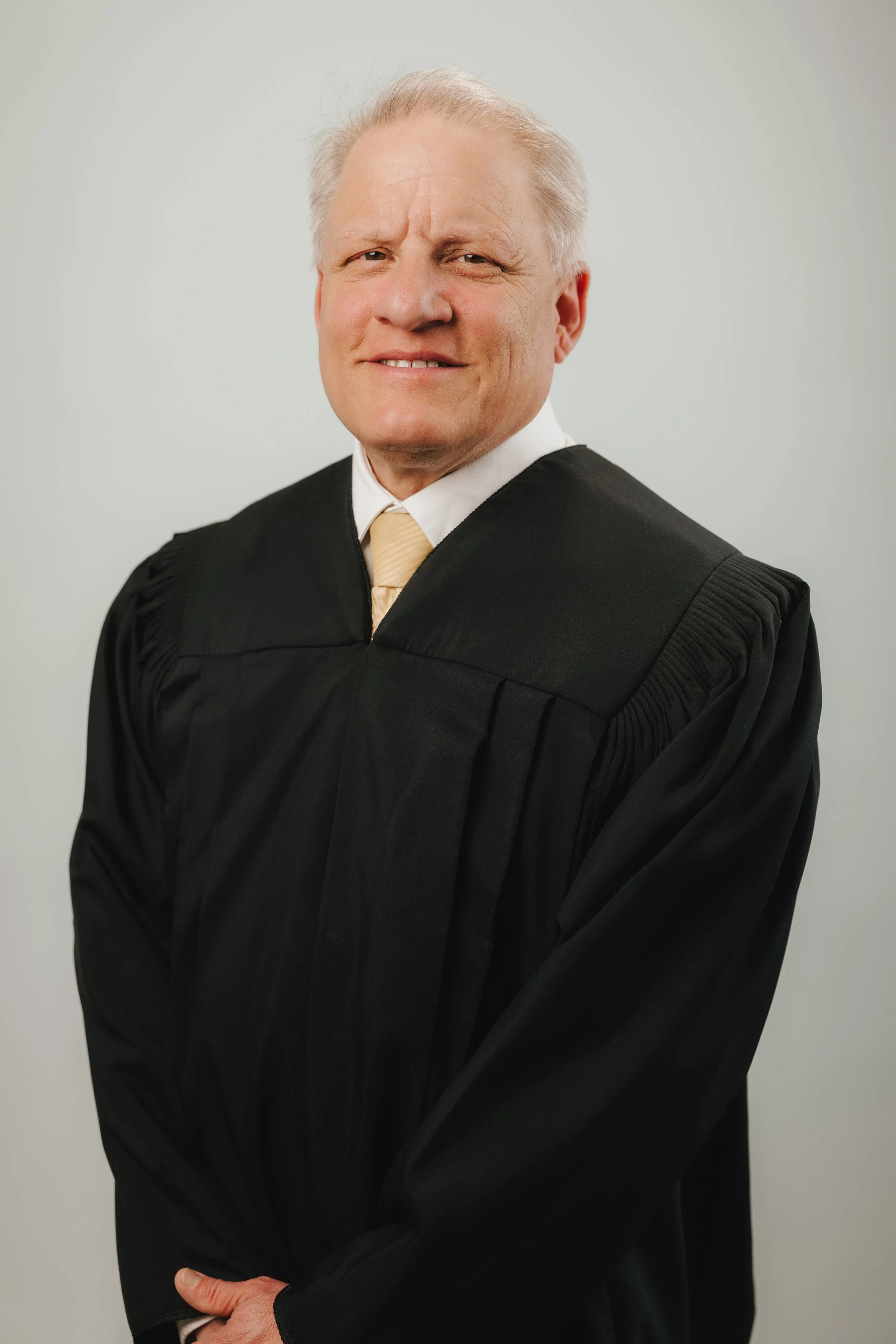 An older man wearing a black judicial robe with a white shirt and light-colored tie, standing against a plain light background.