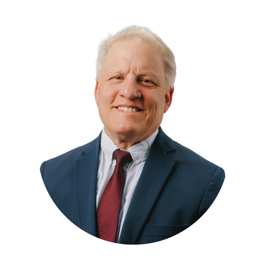 A middle-aged man with light skin, grey hair, and a friendly smile, dressed in a dark blue suit, a light blue shirt, and a maroon tie, posing in front of a plain background.